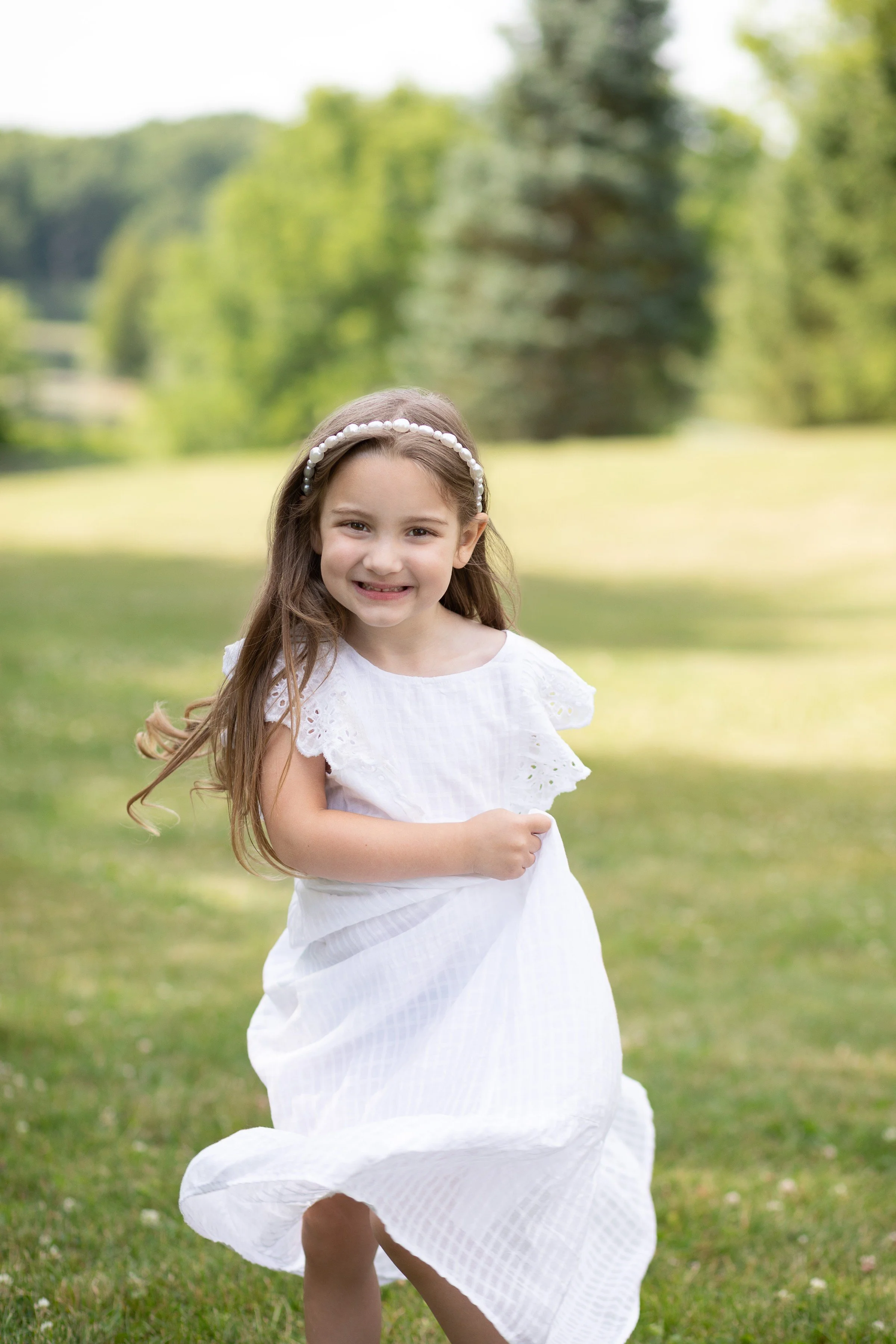 Young girl in a white dress running outdoors in a grassy area with trees in the background.