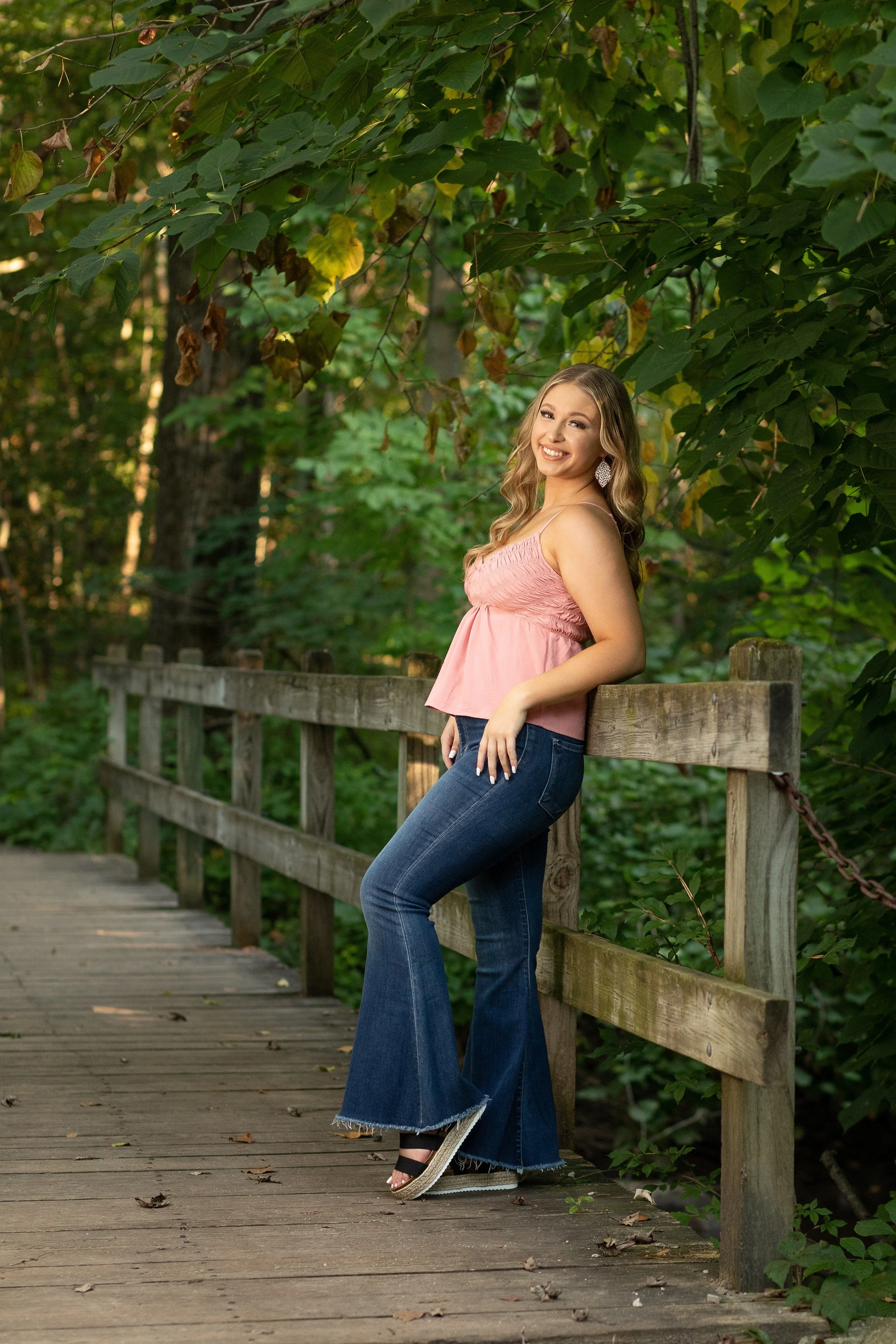 A young woman with wavy blonde hair in a pink sleeveless top and flared jeans standing on a wooden bridge in a lush green forest, smiling at the camera.