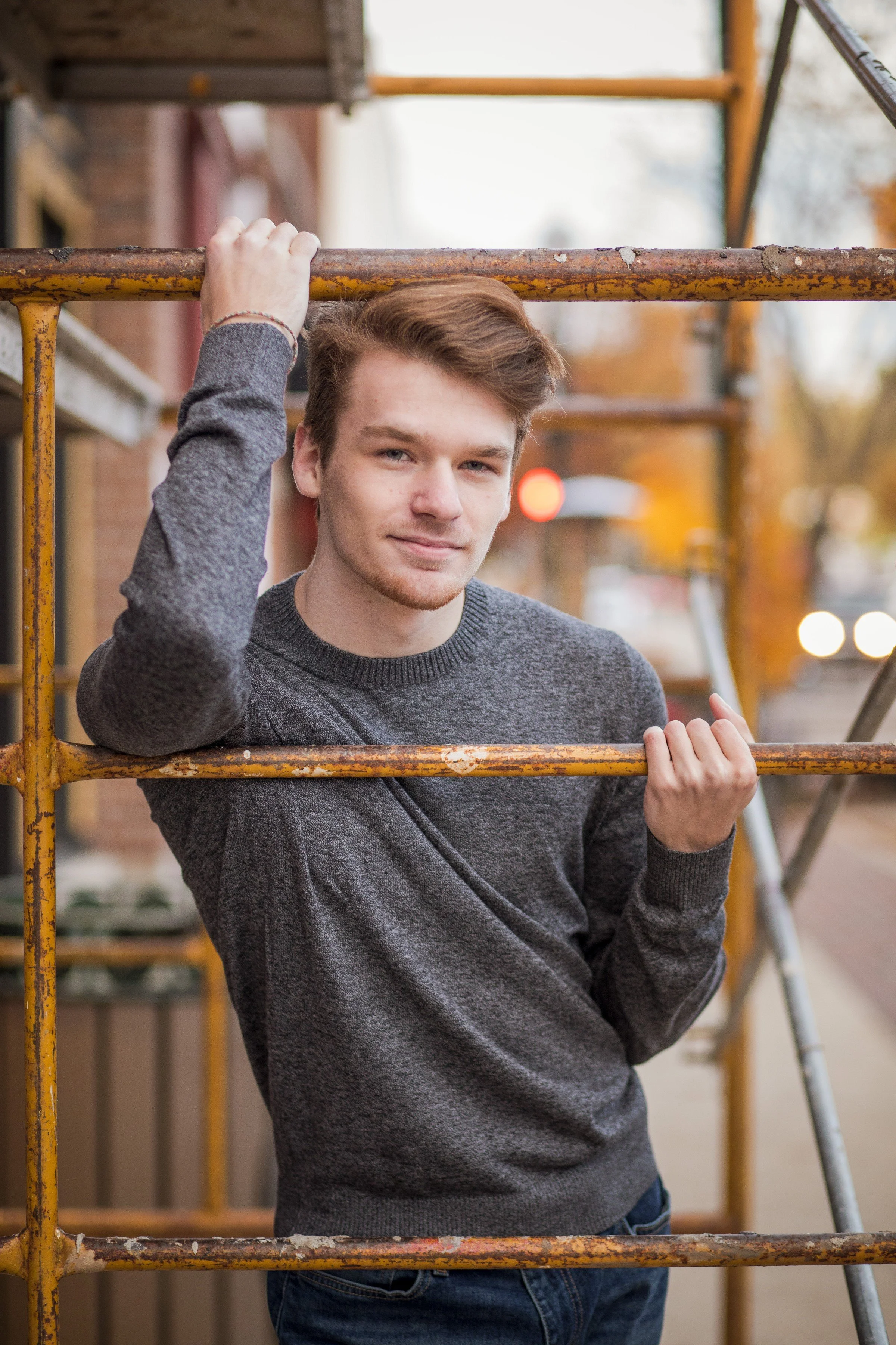 A young man with light brown hair and a grey sweater standing behind yellow metal scaffolding outdoors on a city street.