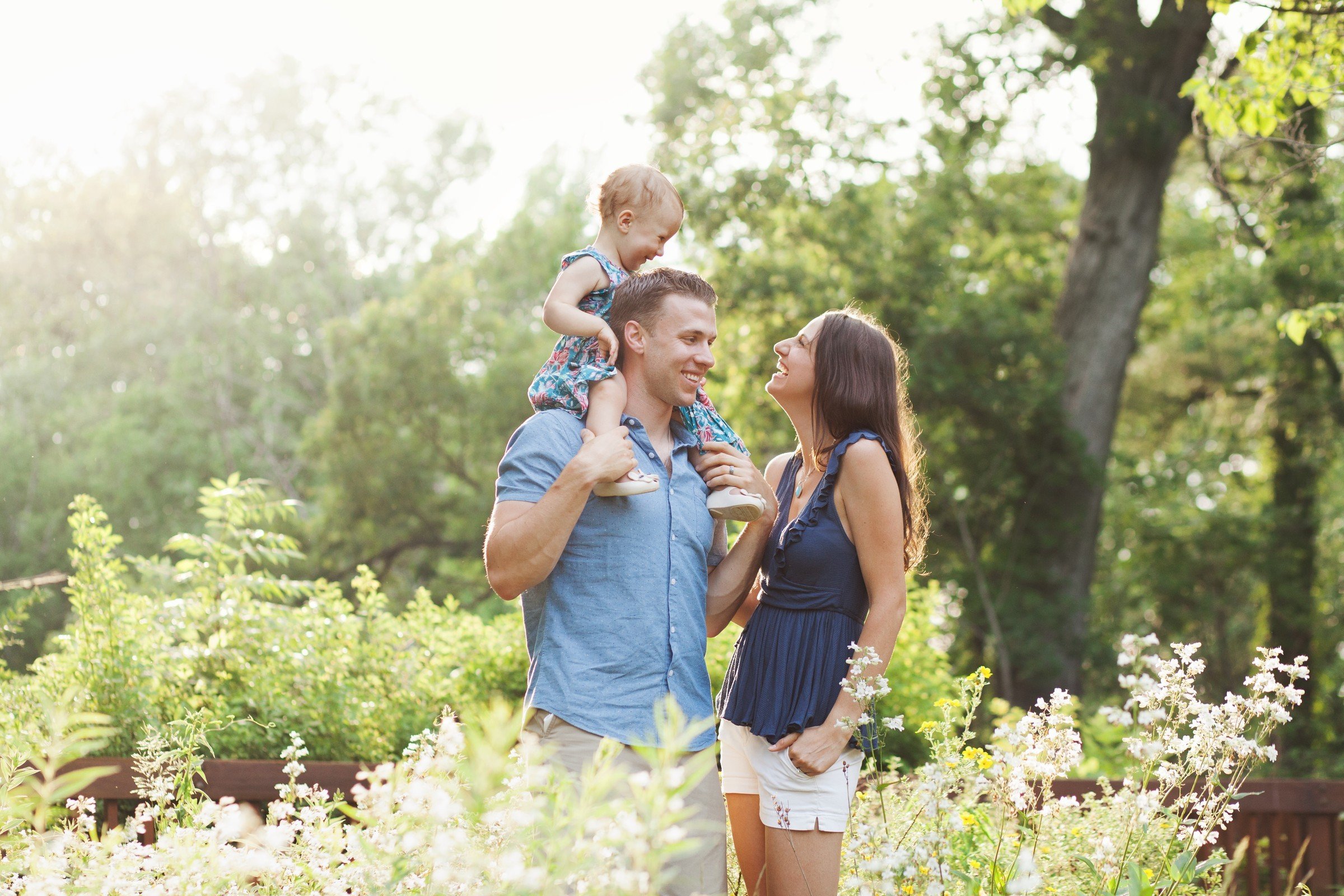 Family of three enjoying a sunny day outdoors, with a man carrying a young girl on his shoulders and a woman standing next to them, all smiling and looking at each other.
