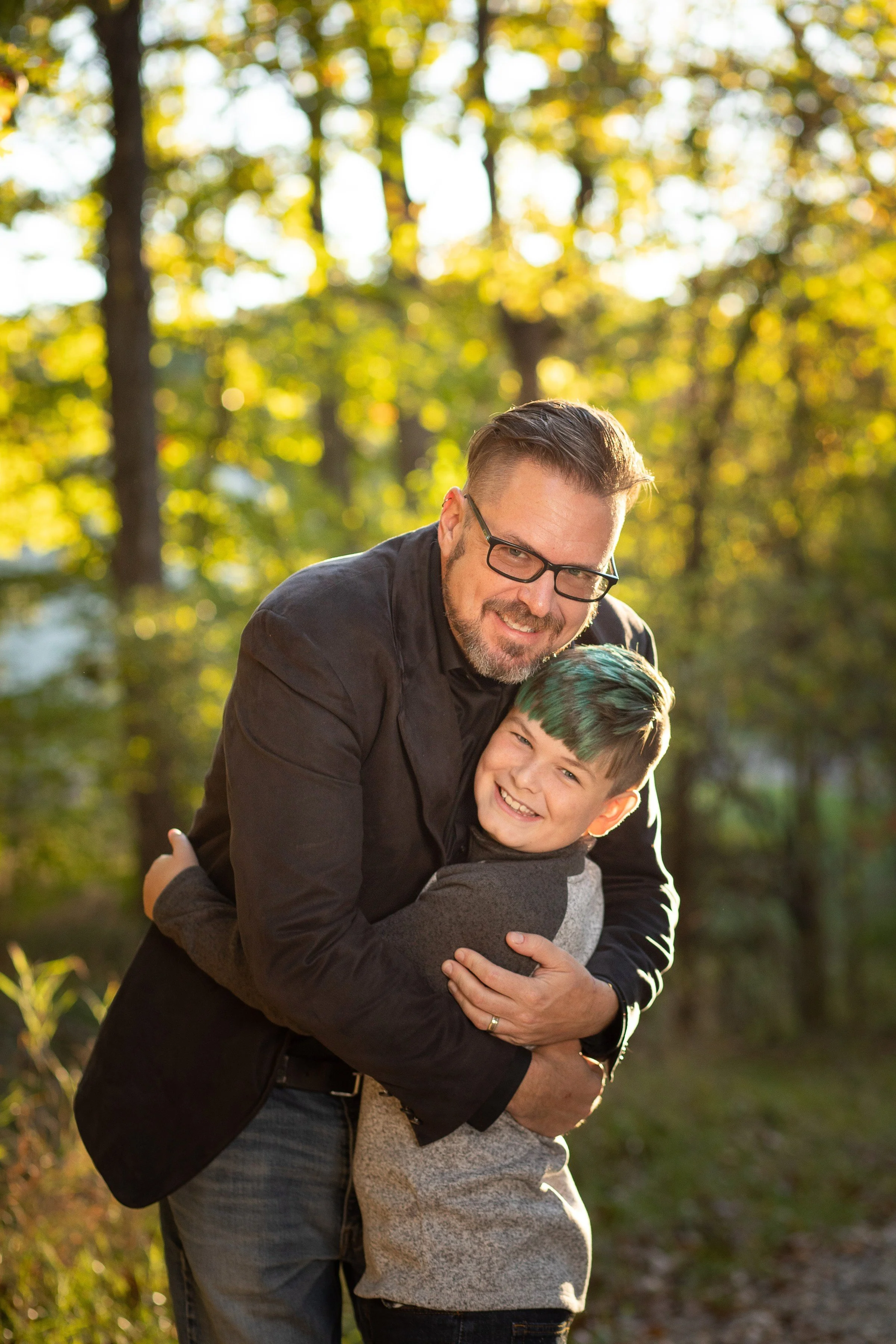 A man hugging a boy outdoors in a forest during sunset, both smiling.