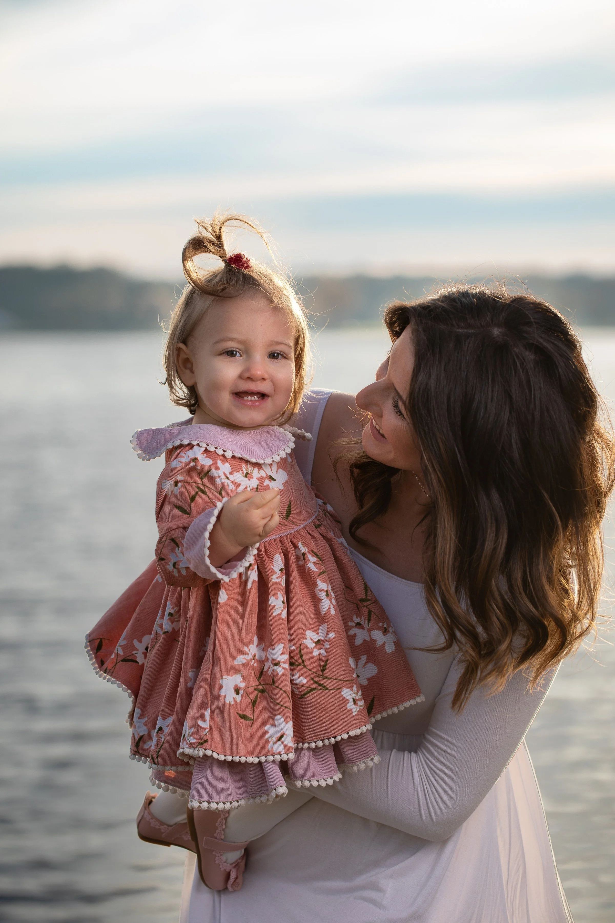 A woman holding a joyful young girl outdoors near a body of water during sunset.