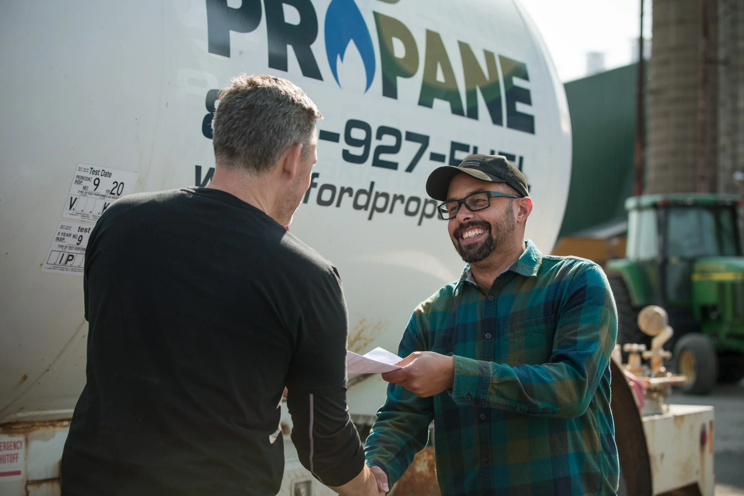 Two men shaking hands outdoors next to propane tanks, one wearing a black shirt and the other wearing a green plaid shirt and glasses, both smiling.