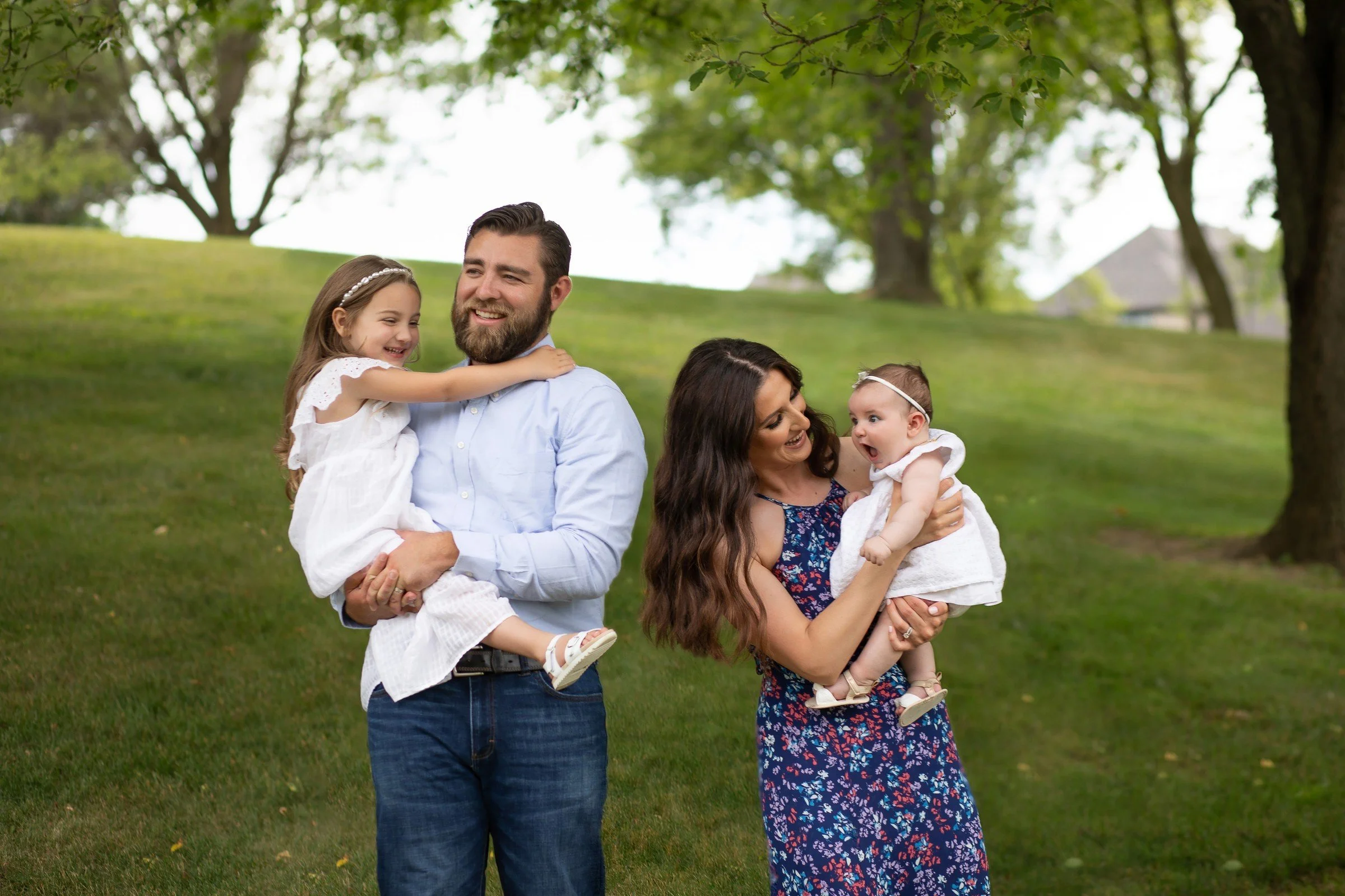 A family with a father, mother, and two young girls standing outdoors on grass with trees in the background. The father is holding the older girl, who is smiling and hugging him. The mother is holding the younger girl, who has a surprised expression.