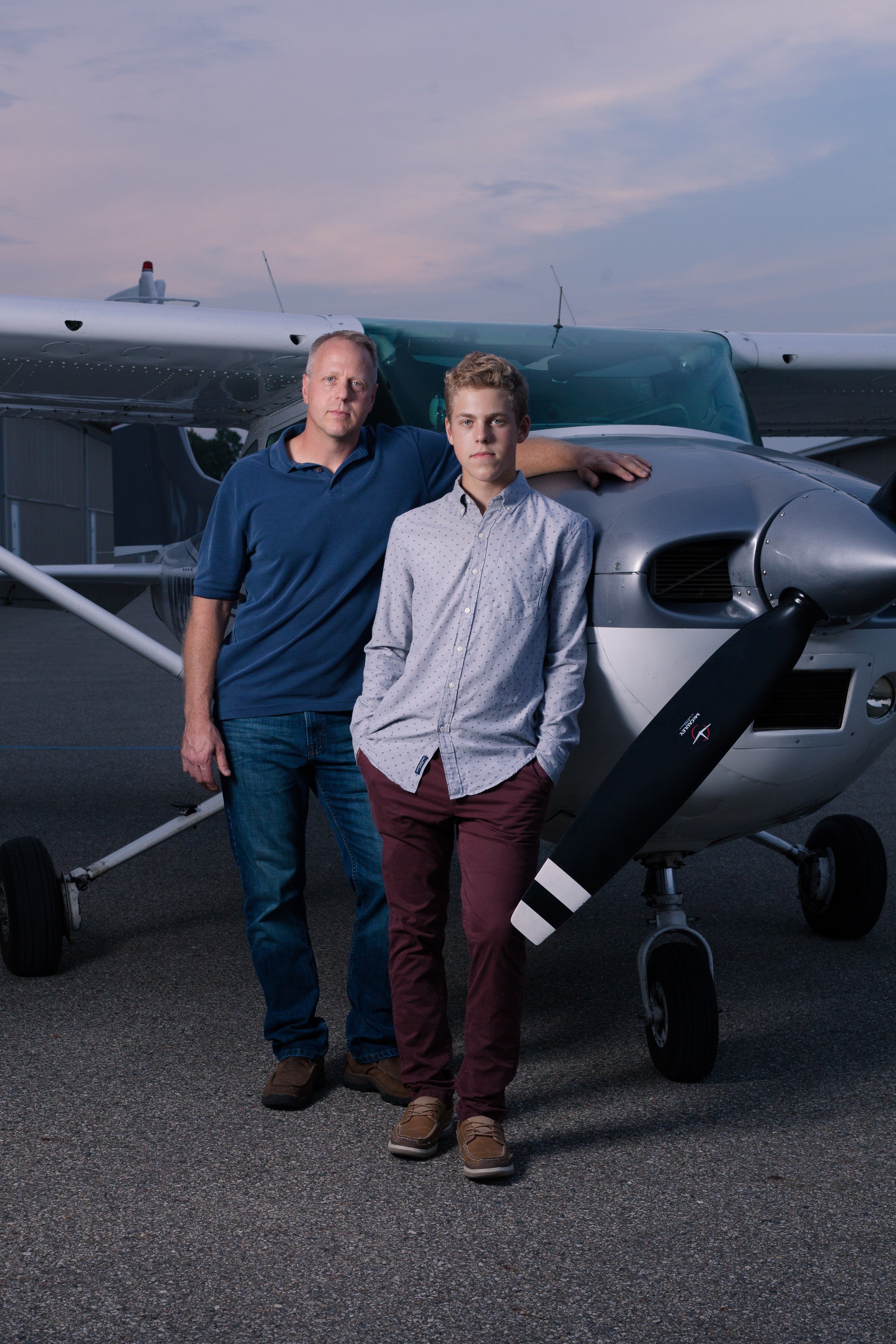 Two males, an adult and a teenager, standing in front of a small airplane during twilight. The adult has his arm resting on the airplane's wing, and both are looking at the camera.