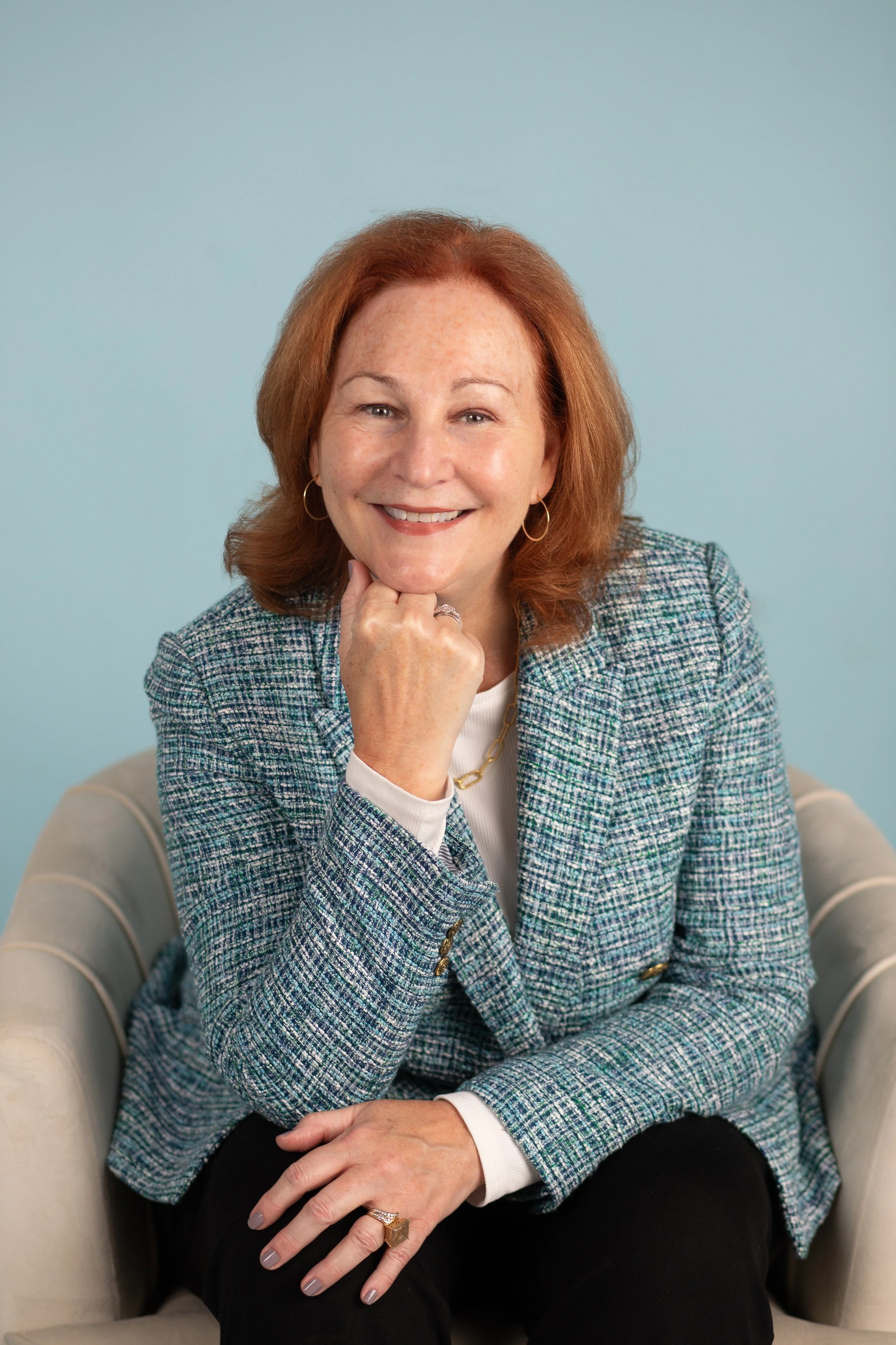 Smiling woman with red hair sitting in a beige chair against a light blue background, wearing a checkered blazer and gold jewelry.
