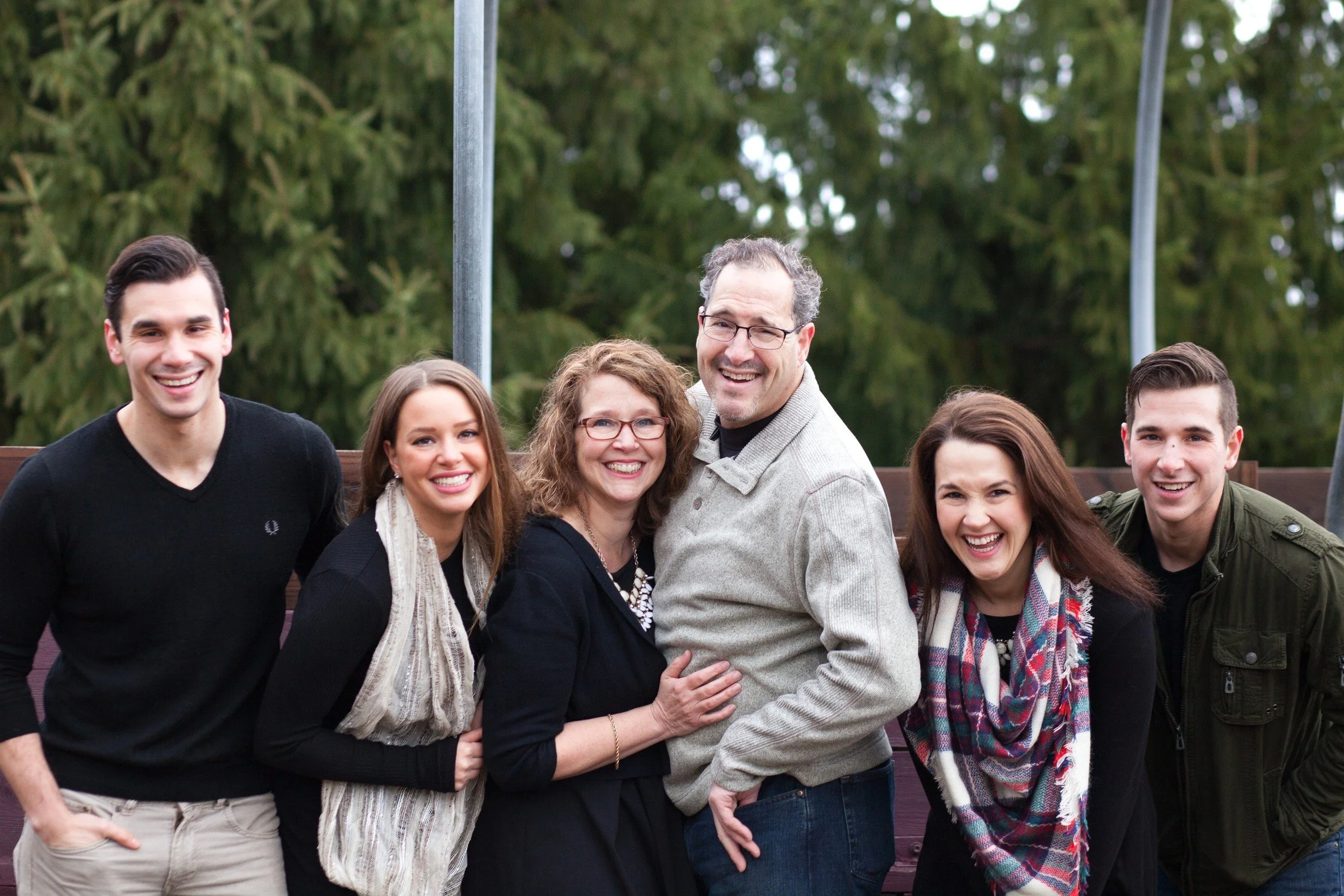 Group of six smiling people, three men and three women, standing outdoors with trees in the background.