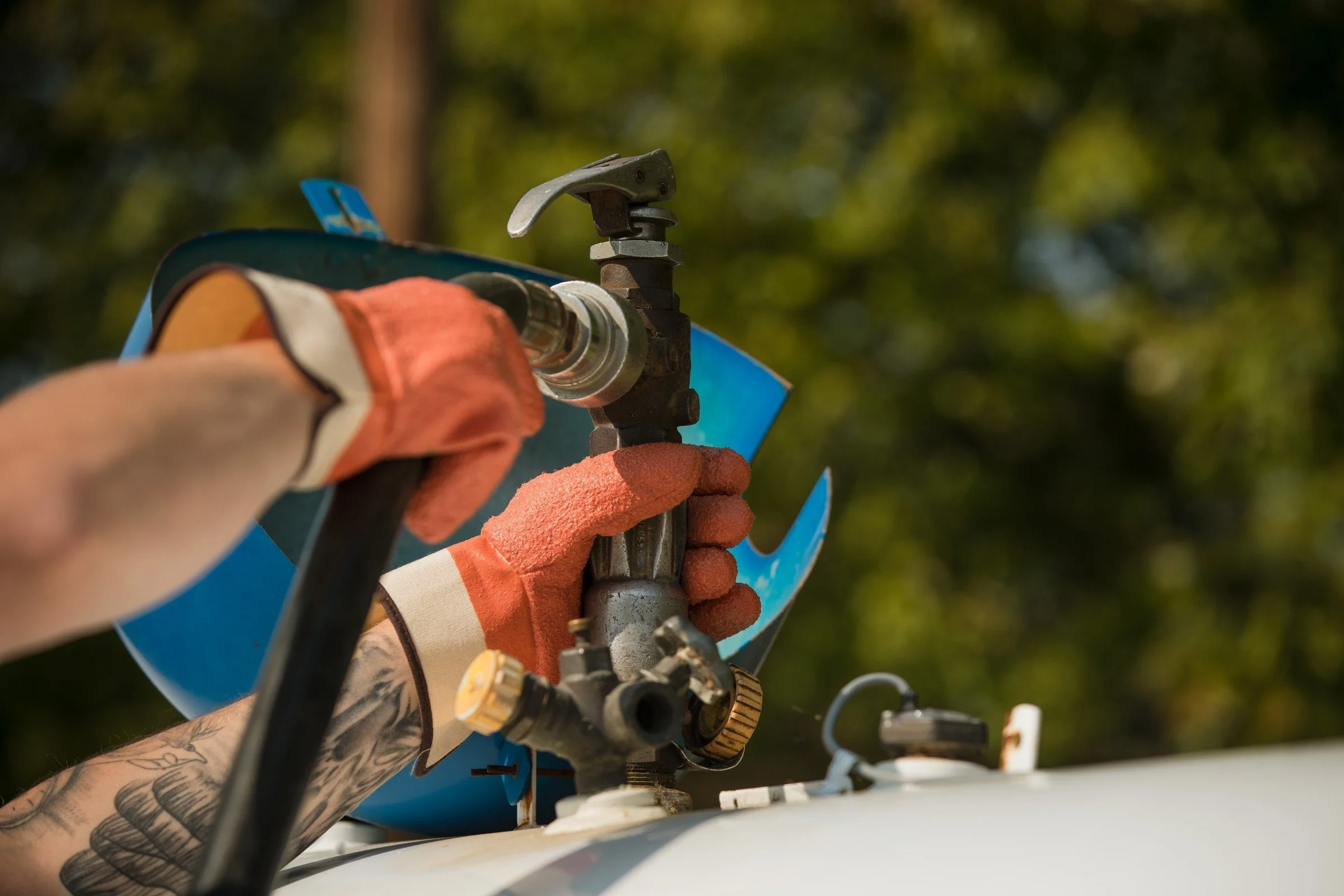 Close-up of a person wearing orange gloves using a tool on a device outdoors with a background of trees.