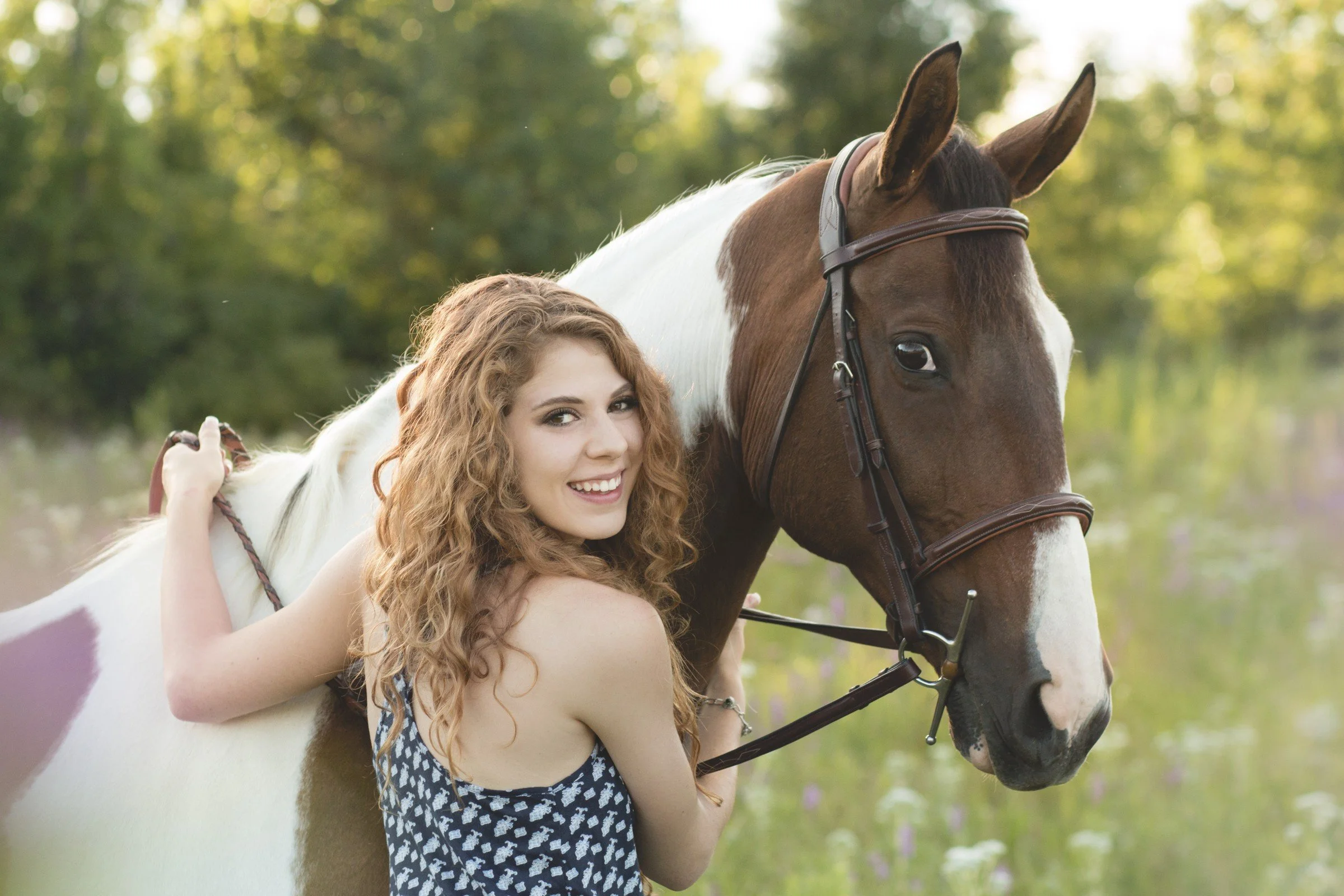 A young woman with curly red hair smiling while hugging a horse with a brown and white coat in a lush green outdoor setting.