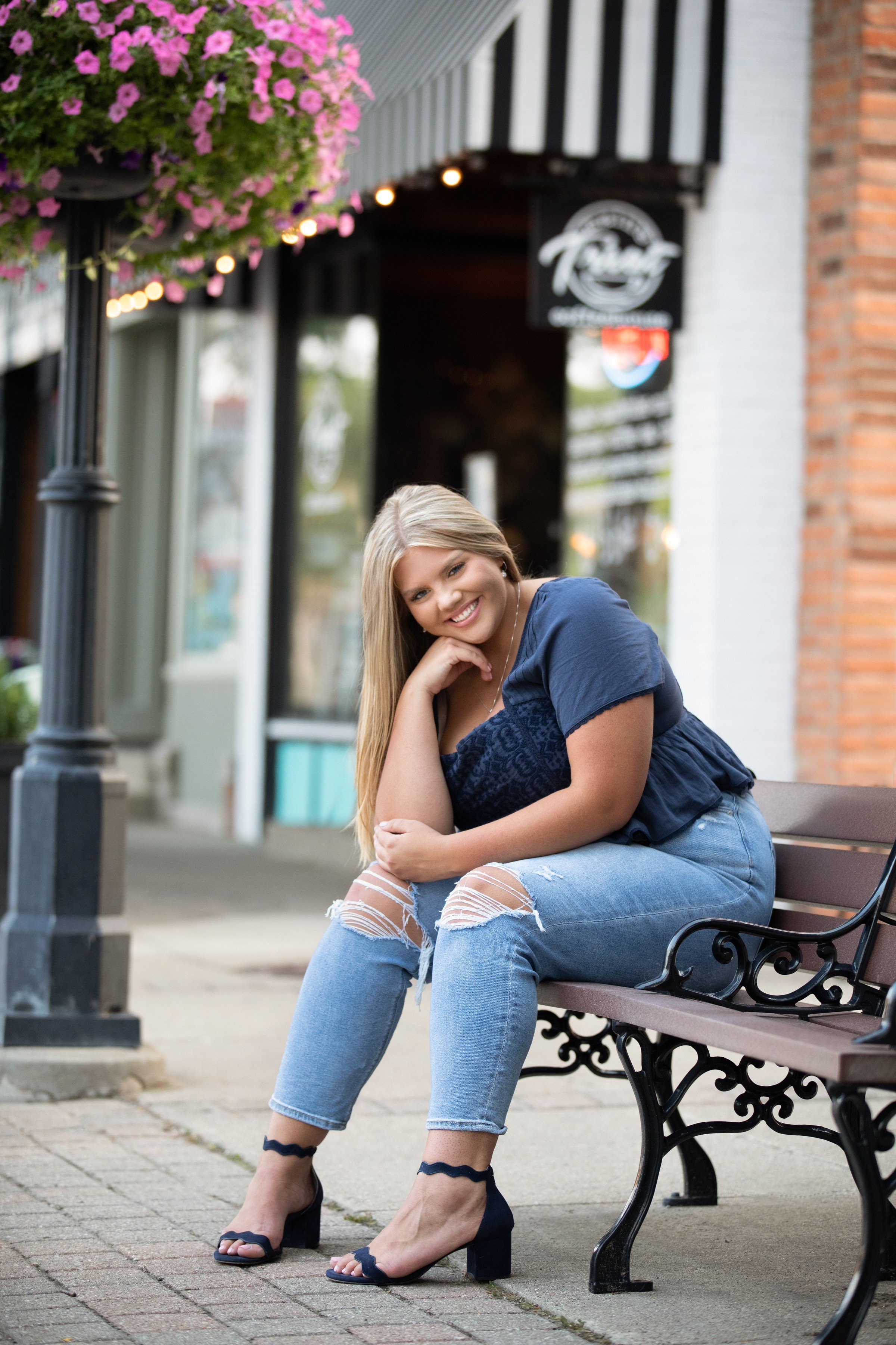 A young woman with blonde hair, wearing a navy top and ripped jeans, sitting on a bench outside a storefront with pink flowers hanging above and smiling at the camera.