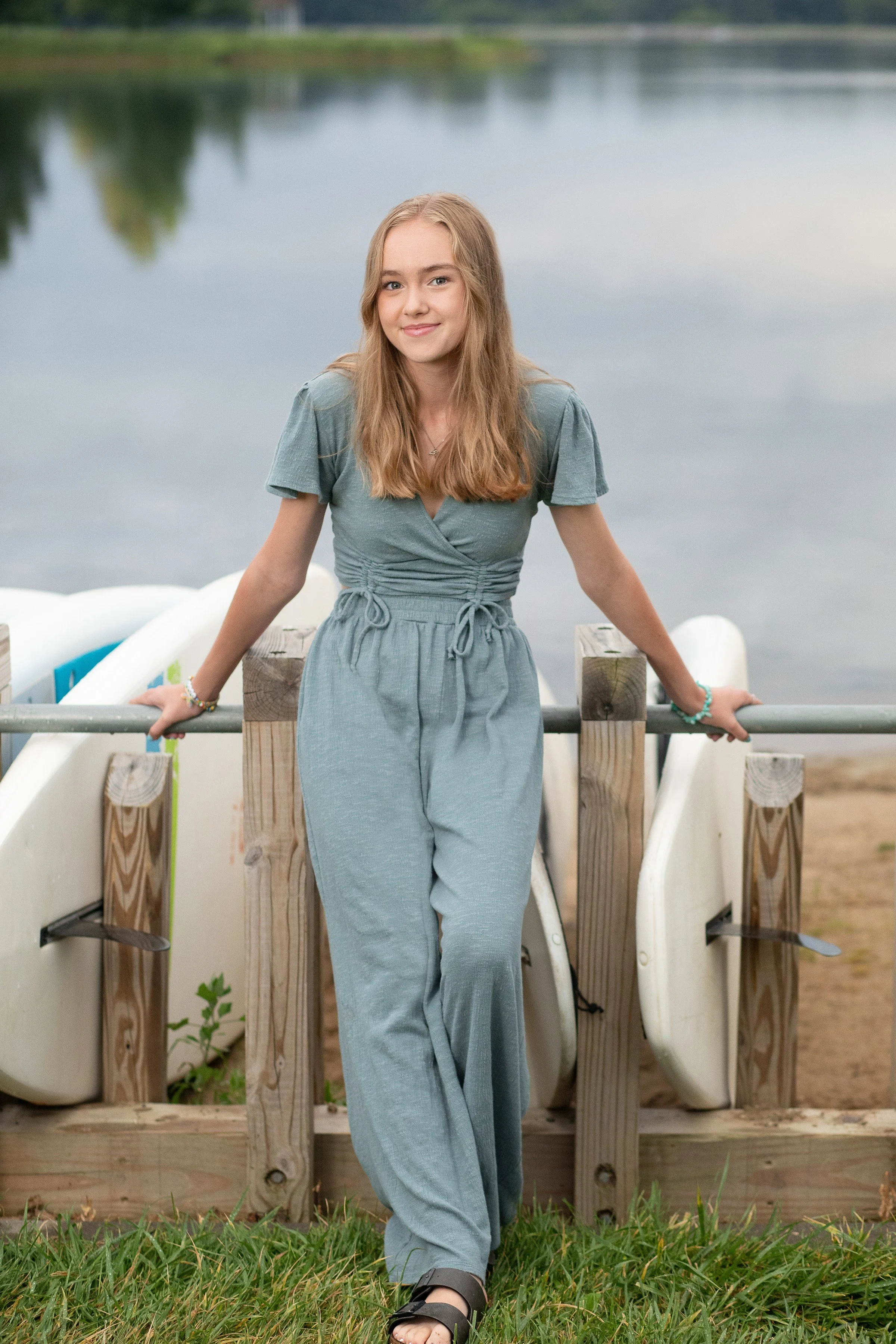 A young woman in a grey outfit standing on grass next to a railing with paddleboards behind her near a lake.
