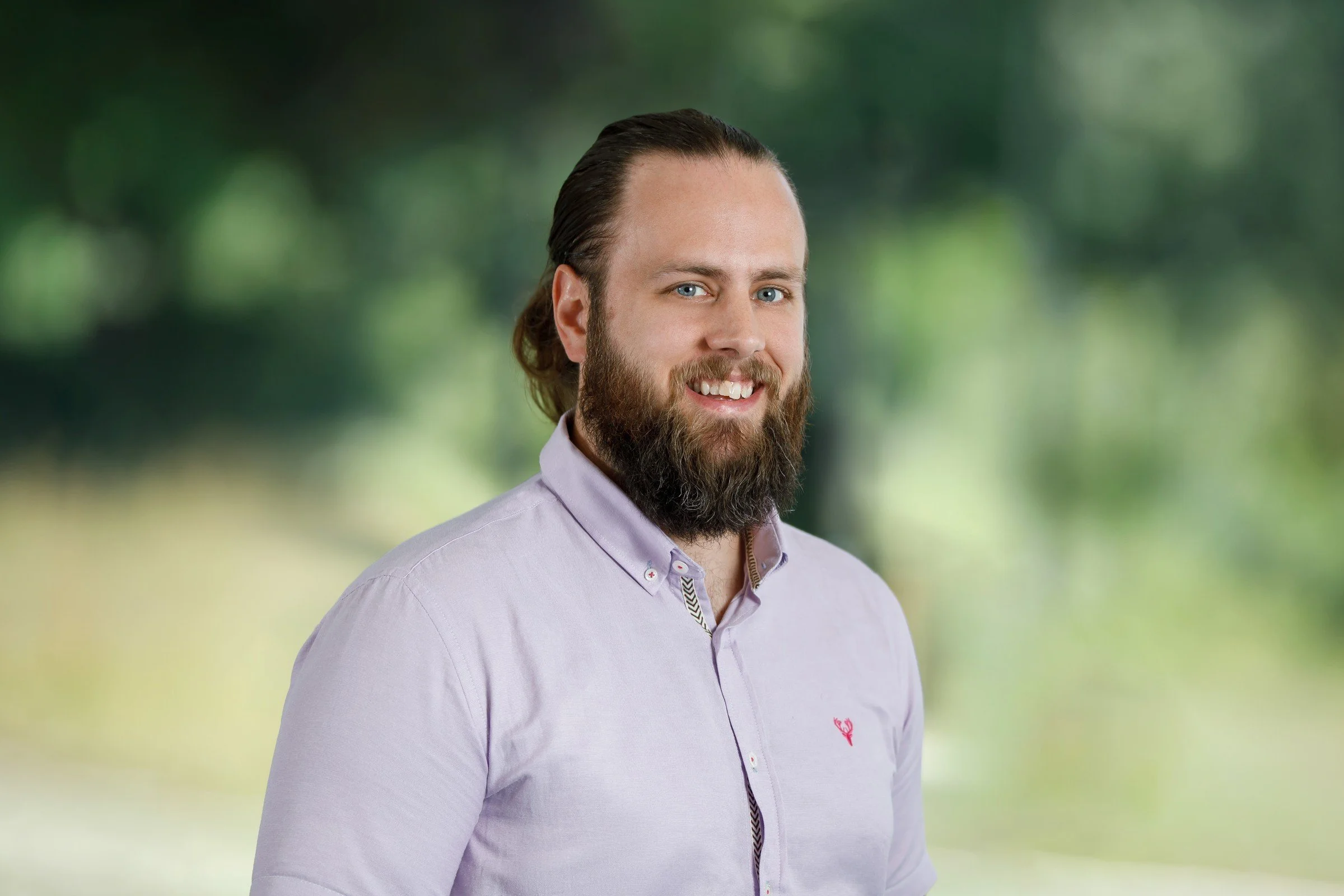 A man with a full beard and long hair, wearing a light purple button-up shirt, smiling against a blurred outdoor background with trees and greenery.