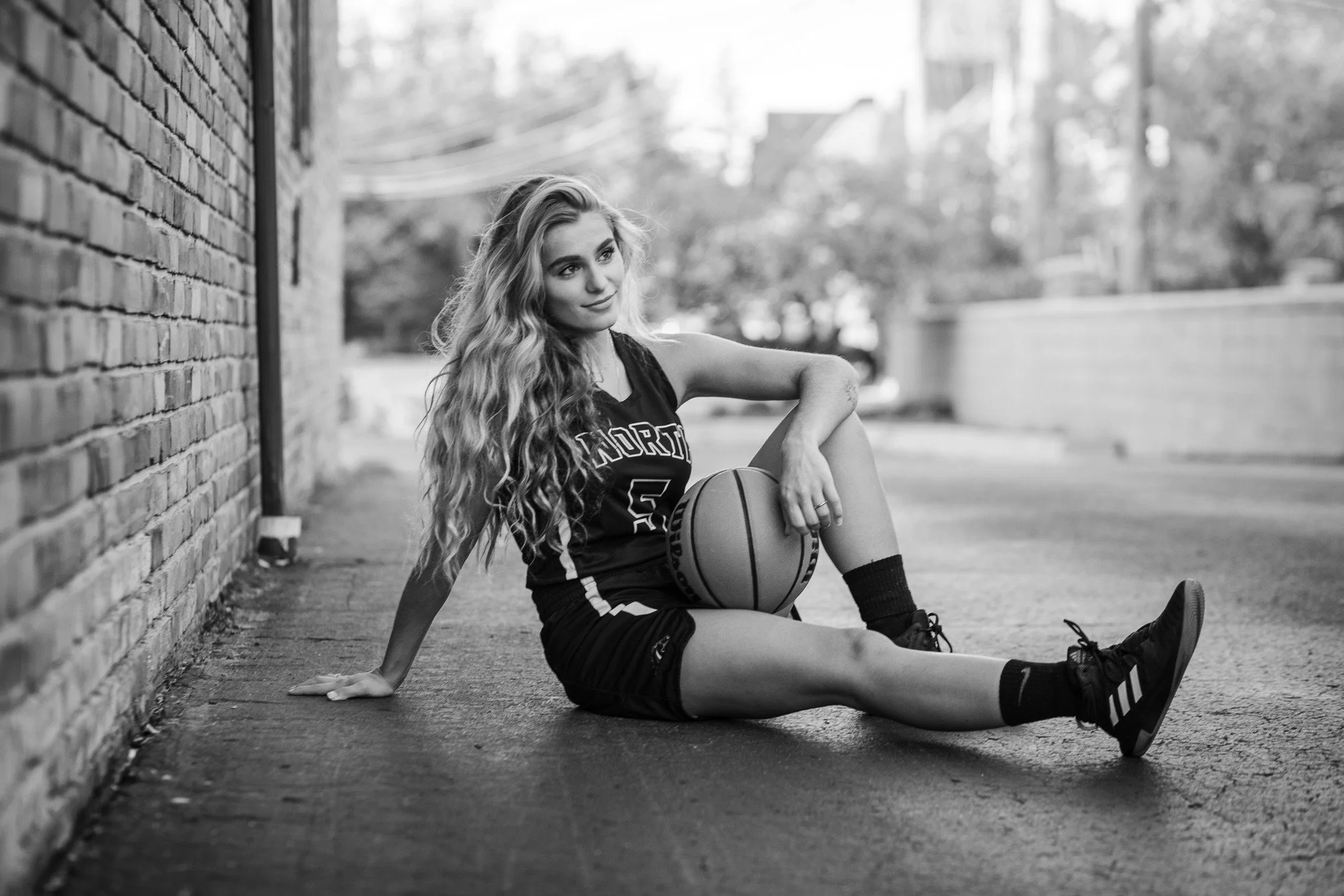 A young woman in a basketball jersey and shorts sitting on the sidewalk, holding a basketball, with one arm resting on the ground and one leg extended. She has long, wavy hair and is wearing sneakers and socks. The background shows a brick wall and t