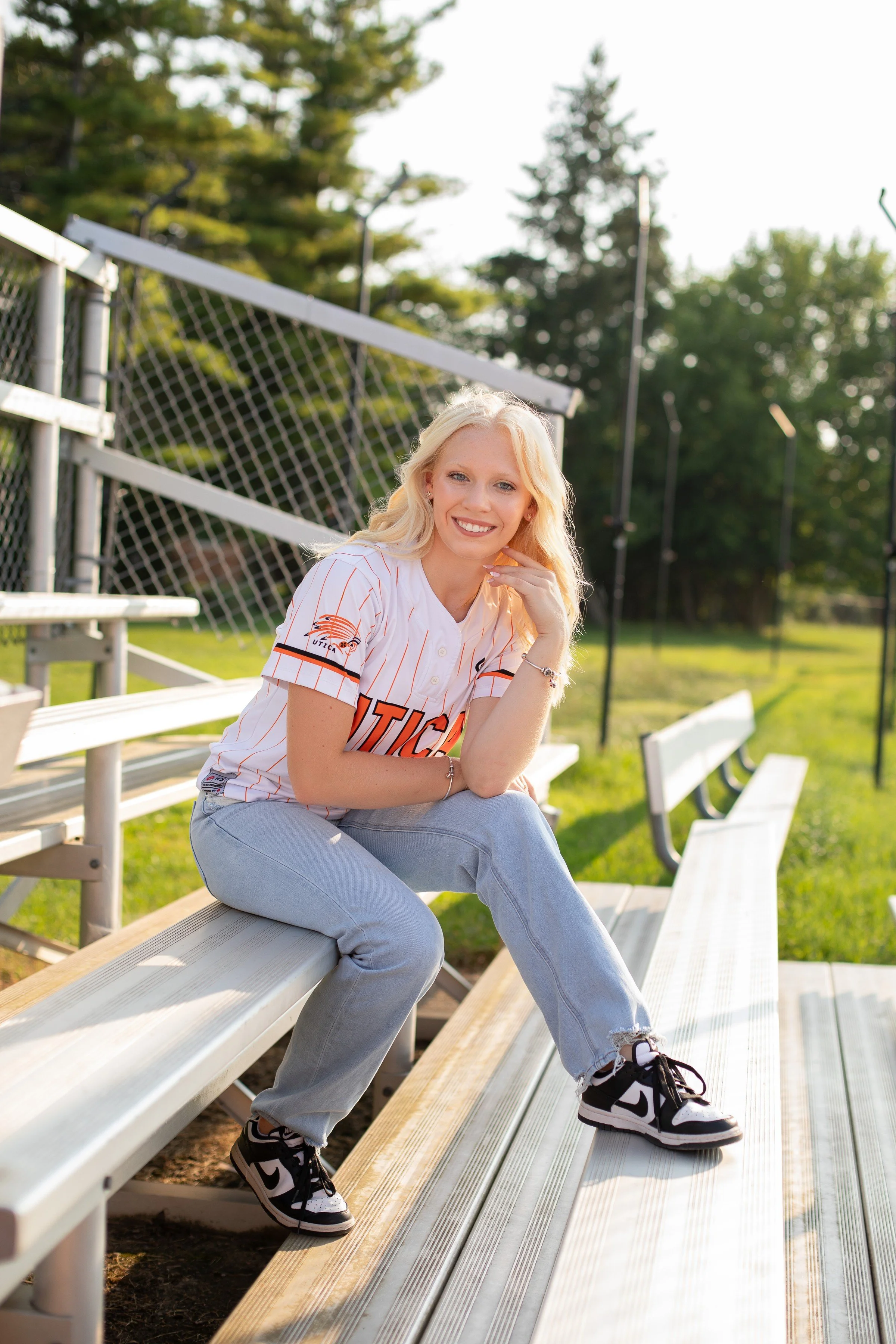 A young woman with blonde hair sitting on bleachers at a sports field, wearing a baseball jersey and sneakers, smiling at the camera during daylight.
