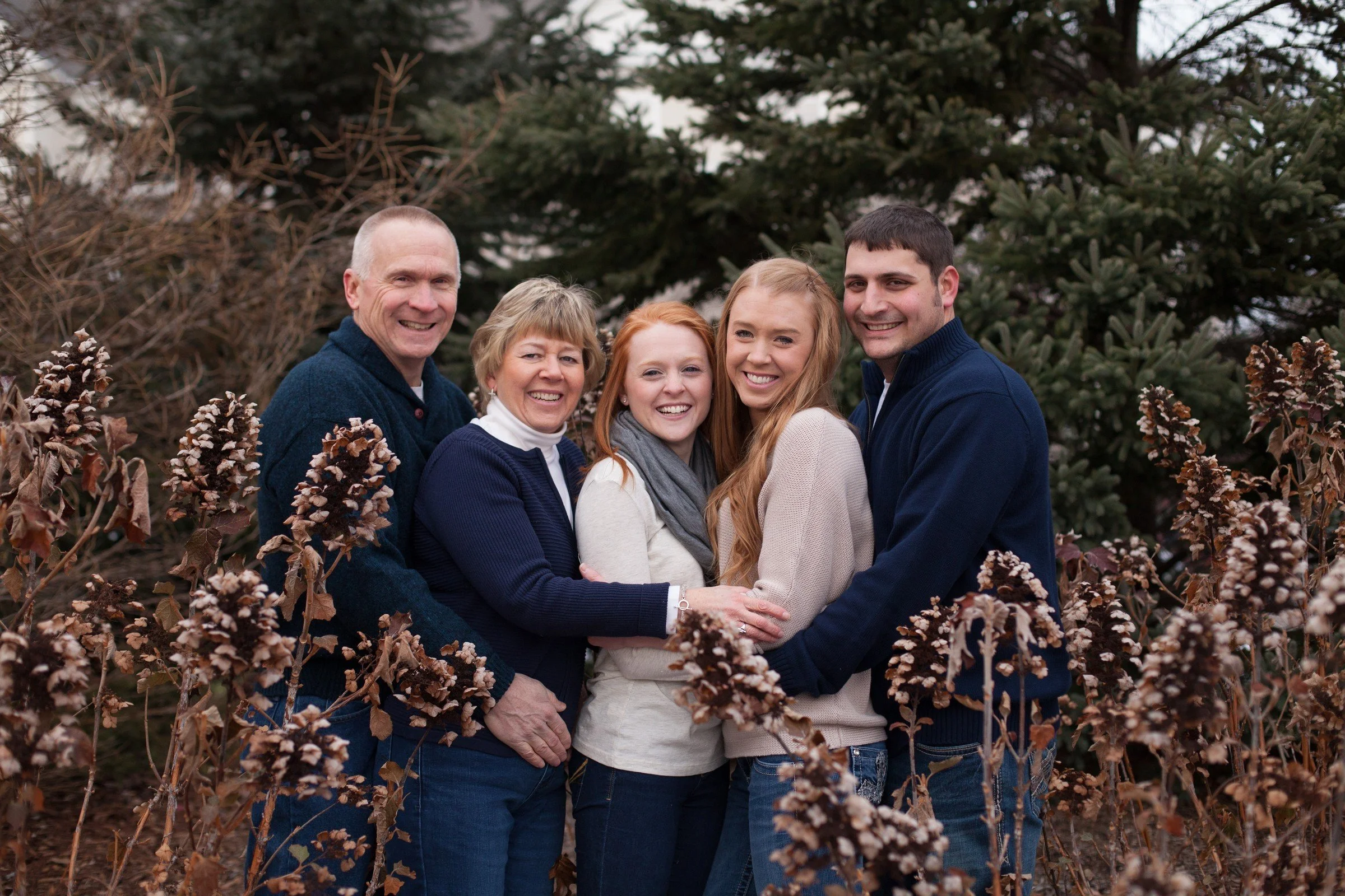 A family of five standing close together outdoors surrounded by brown and green trees, smiling and embracing each other.