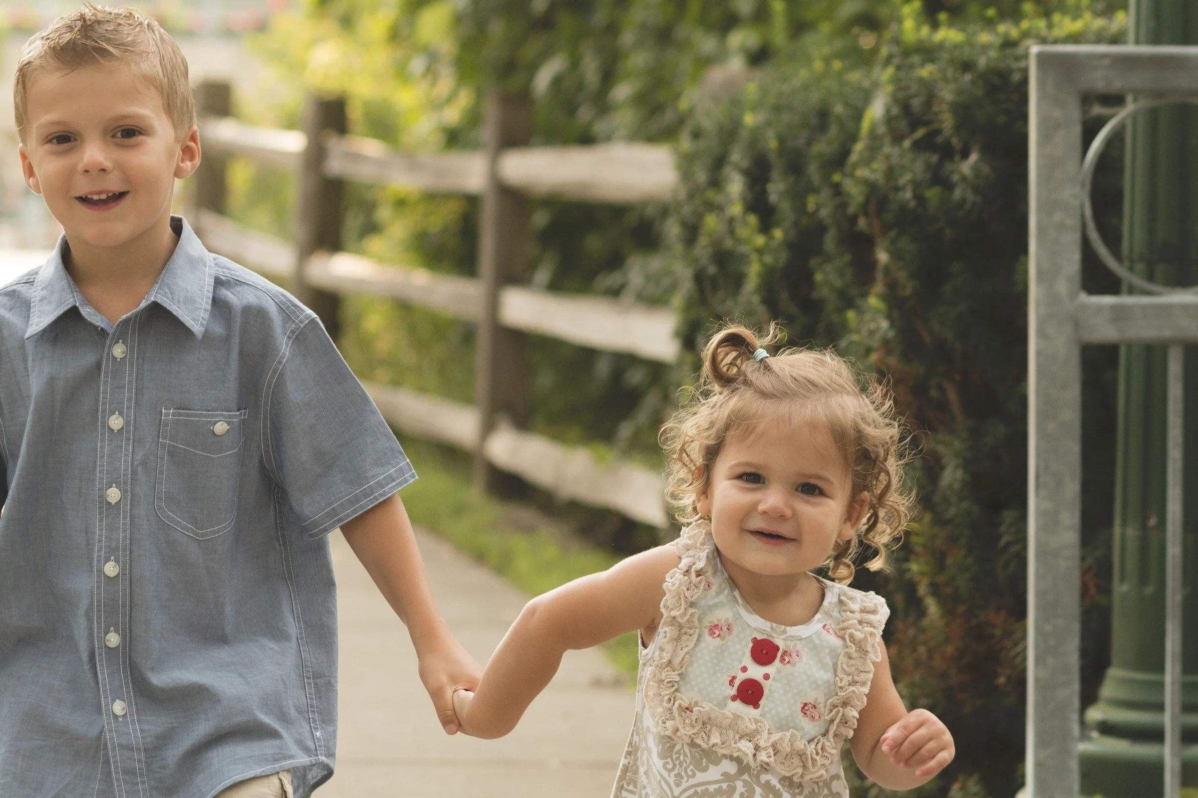 Two young children, a boy and a girl, holding hands and walking outdoors on a sidewalk with greenery and a wooden fence in the background.
