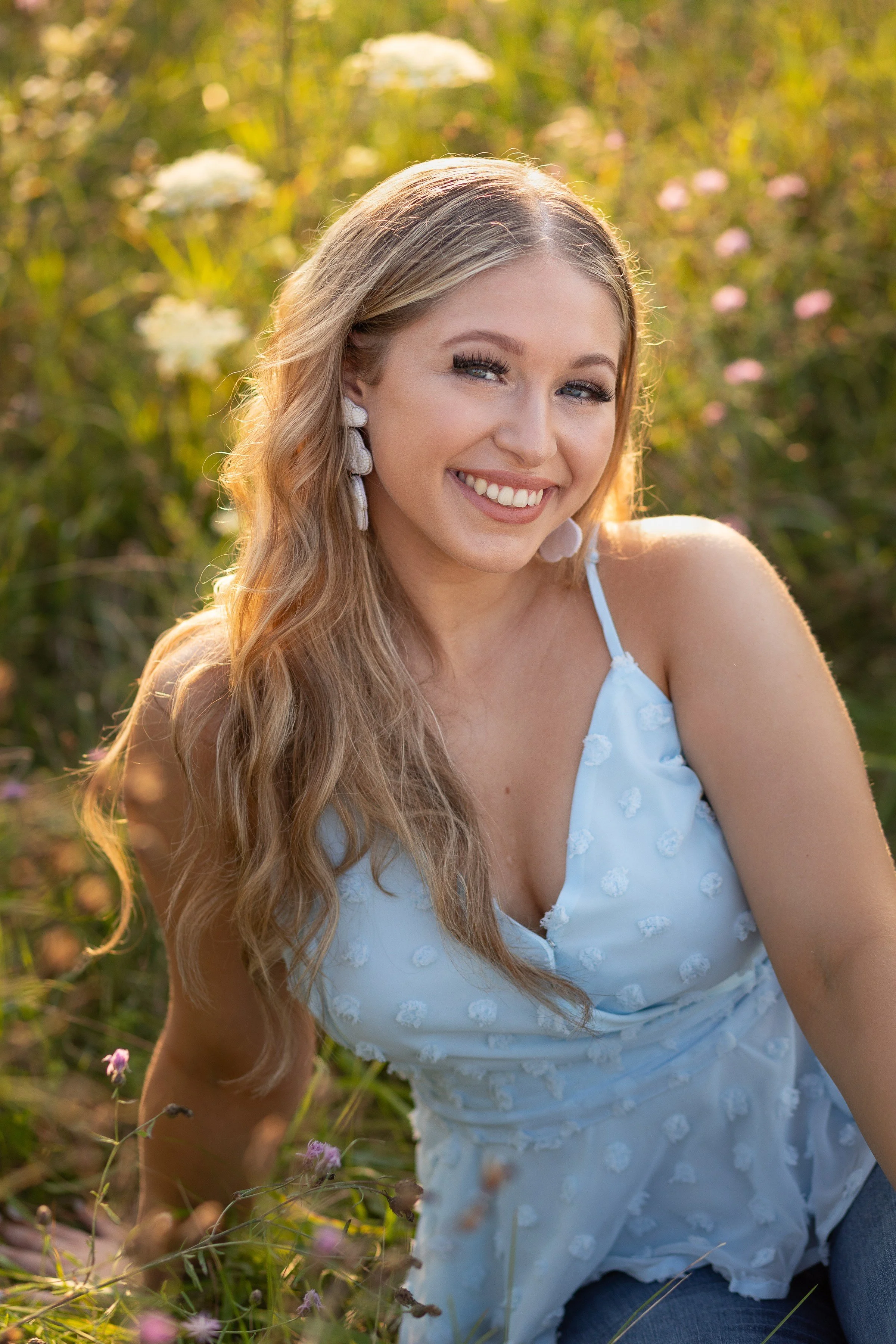 A young woman with long, wavy blonde hair smiling outdoors in a field of wildflowers, wearing a light blue, sleeveless dress with textured patterns.