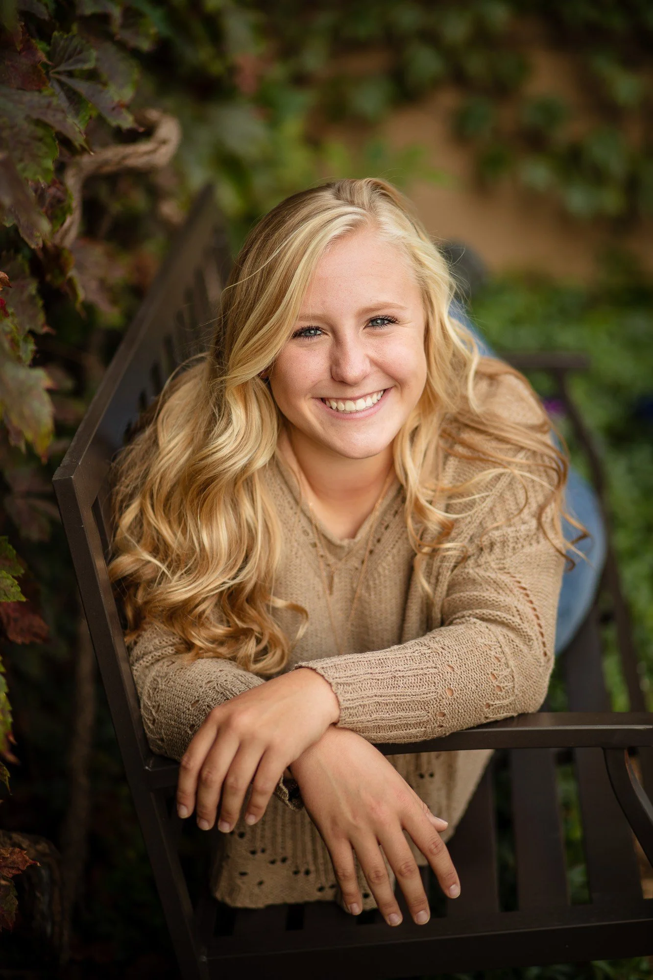 A young blonde woman smiling, sitting on a black bench outdoors with green foliage in the background.