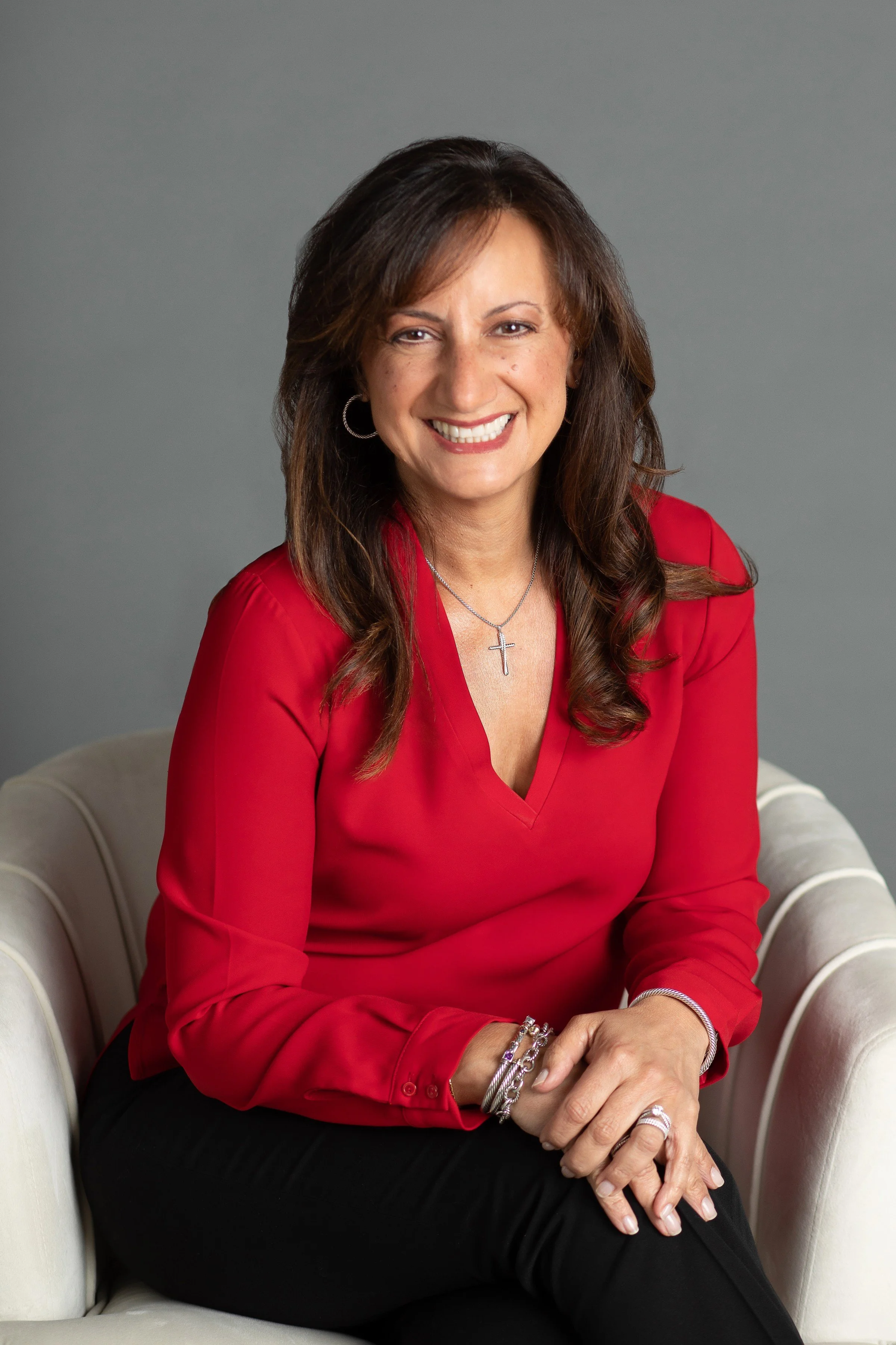 A woman with brown hair and a bright smile, wearing a red blouse and silver jewelry, sitting on a cream-colored armchair against a gray background.