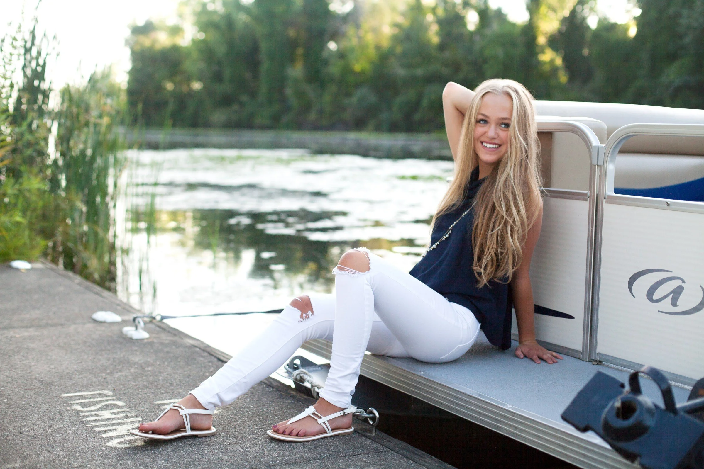 Young woman with long blonde hair wearing a black top and white ripped jeans sitting on the edge of a boat dock near a river, smiling and looking at the camera with one hand behind her head, surrounded by green trees.