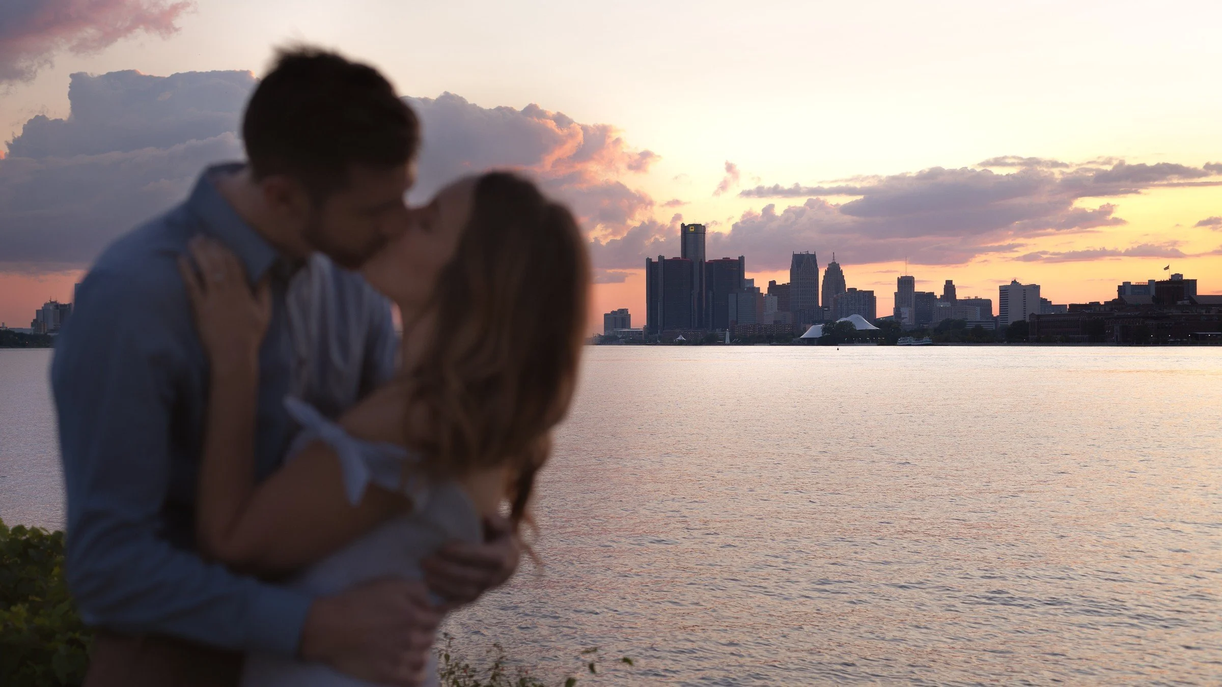 A romantic couple kissing by a body of water during sunset with a city skyline in the background.