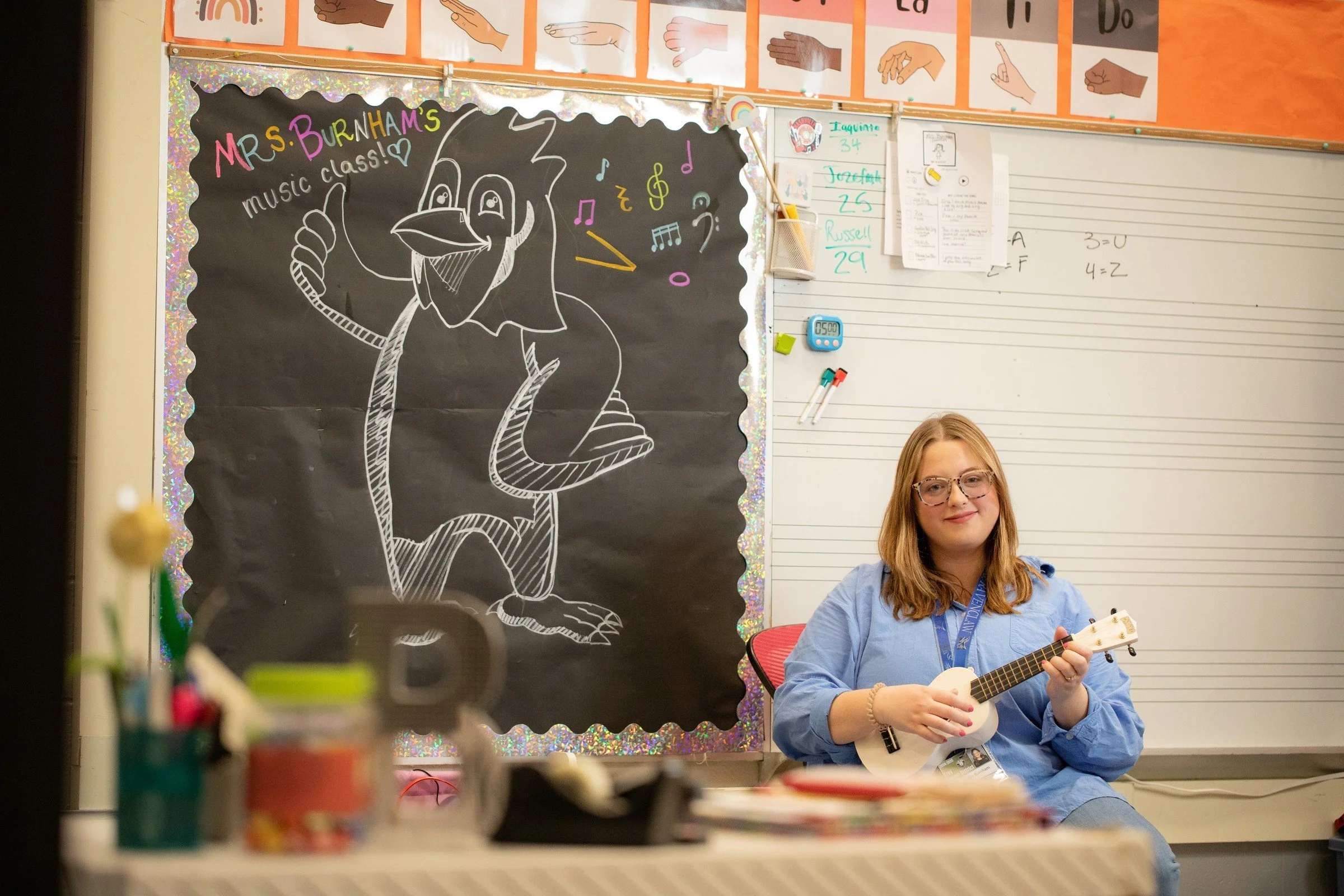 A teacher or student sitting and playing ukulele in front of a classroom decorated with a large black poster featuring a cartoon bird and colorful musical notes. The classroom has educational posters, whiteboard, and supplies.