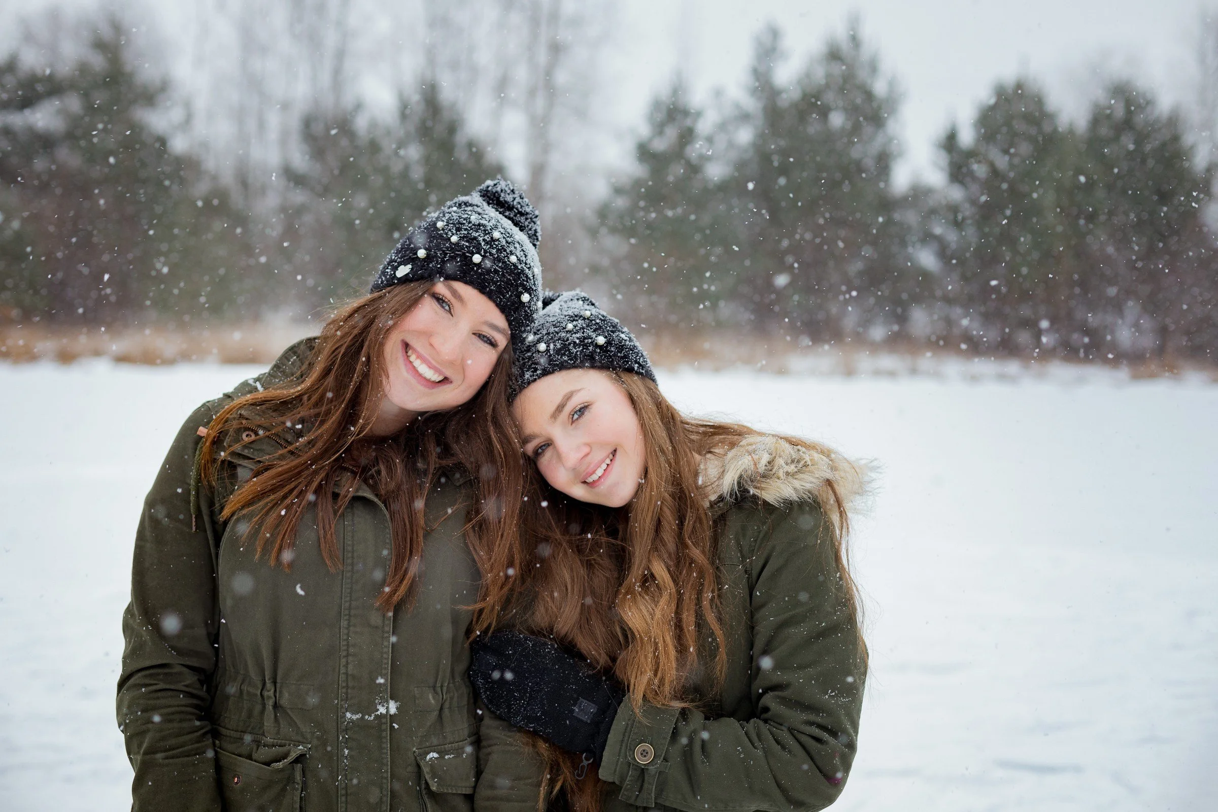Two smiling young women wearing winter coats and knit hats, standing close together in a snowy outdoor setting with snow falling around them.