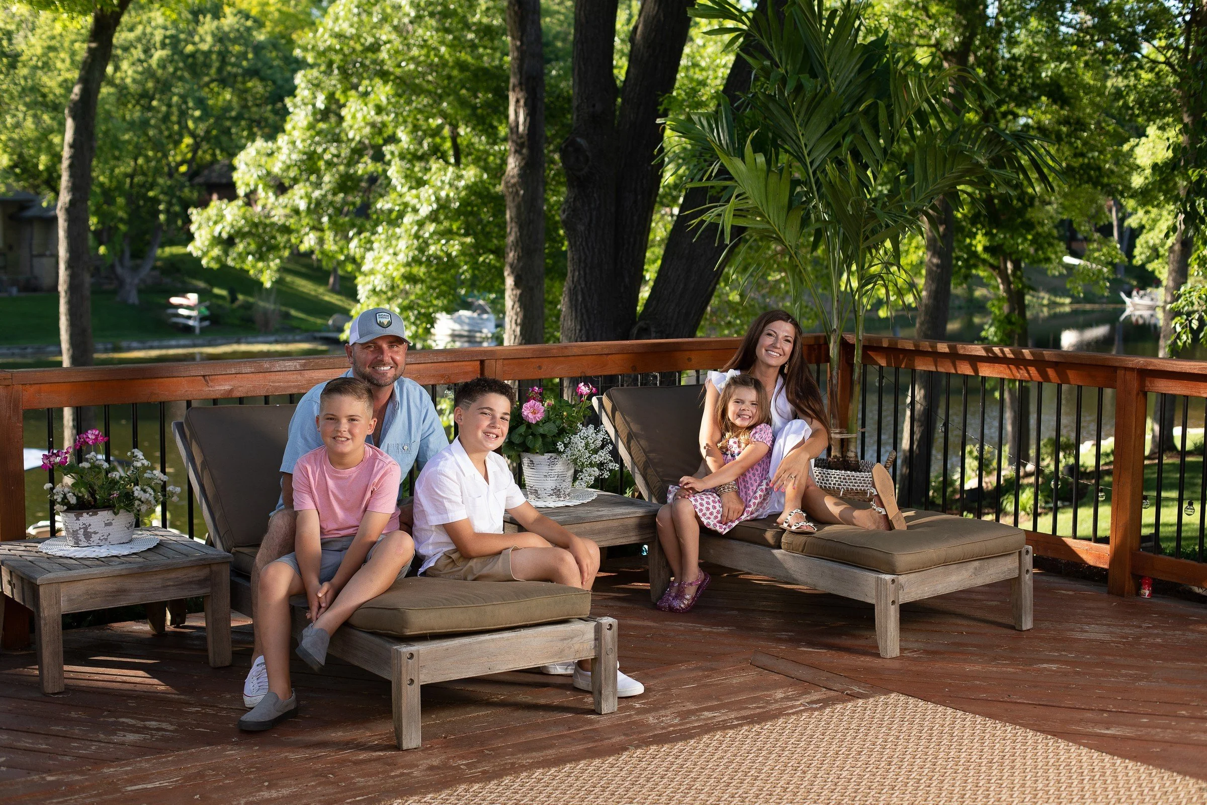Happy family of six sitting and relaxing on a wooden deck overlooking a lush green yard and a river, with trees and flowering plants surrounding them.