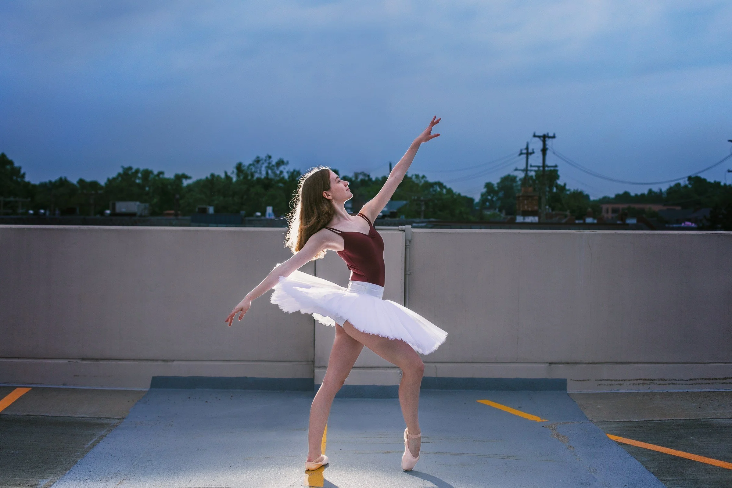 A ballet dancer practices on a rooftop parking lot during dusk, wearing a maroon leotard, white tutu, and pointe shoes, with a cityscape and power lines in the background.