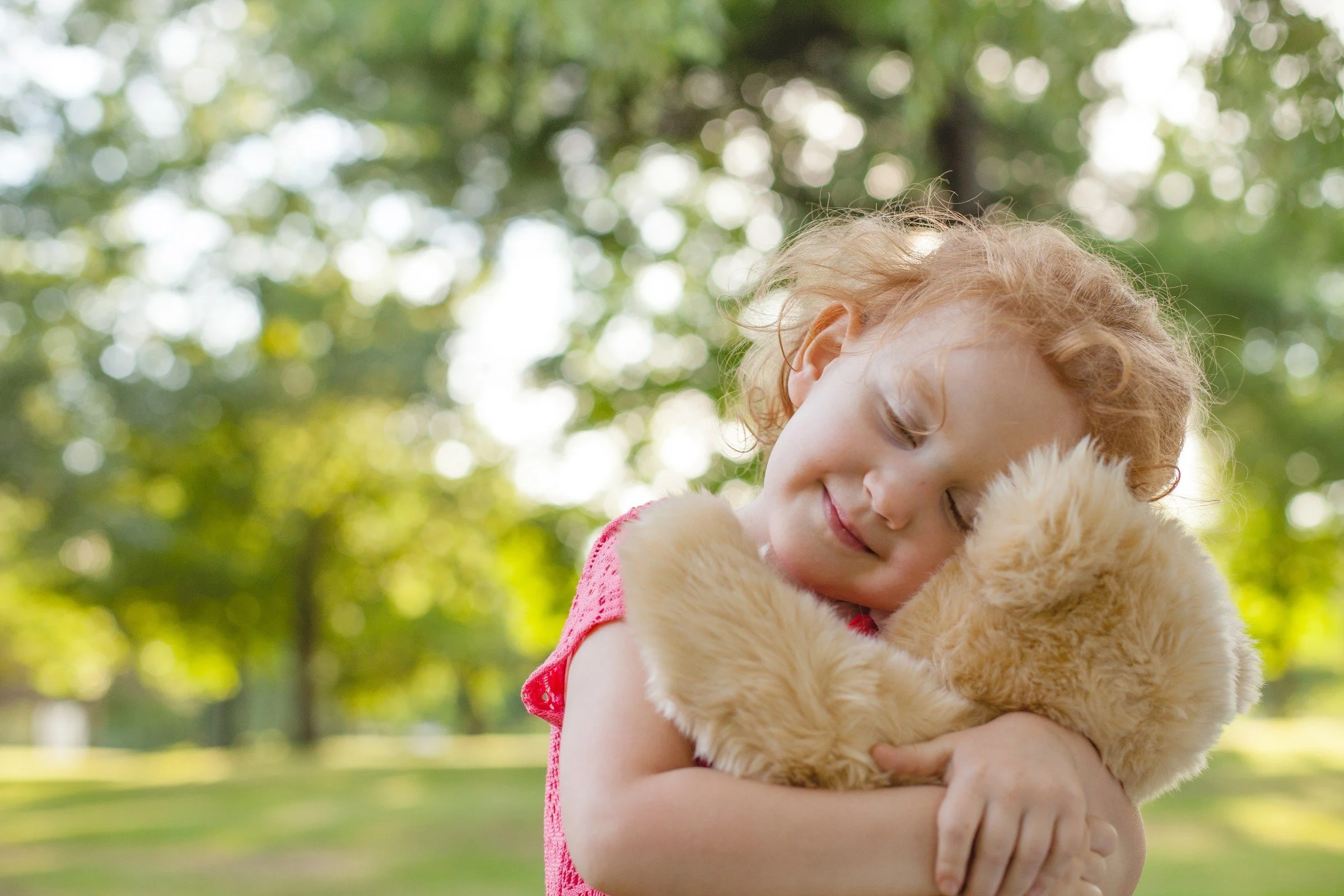 A young girl hugging a teddy bear outdoors on a sunny day, with blurred green trees in the background.