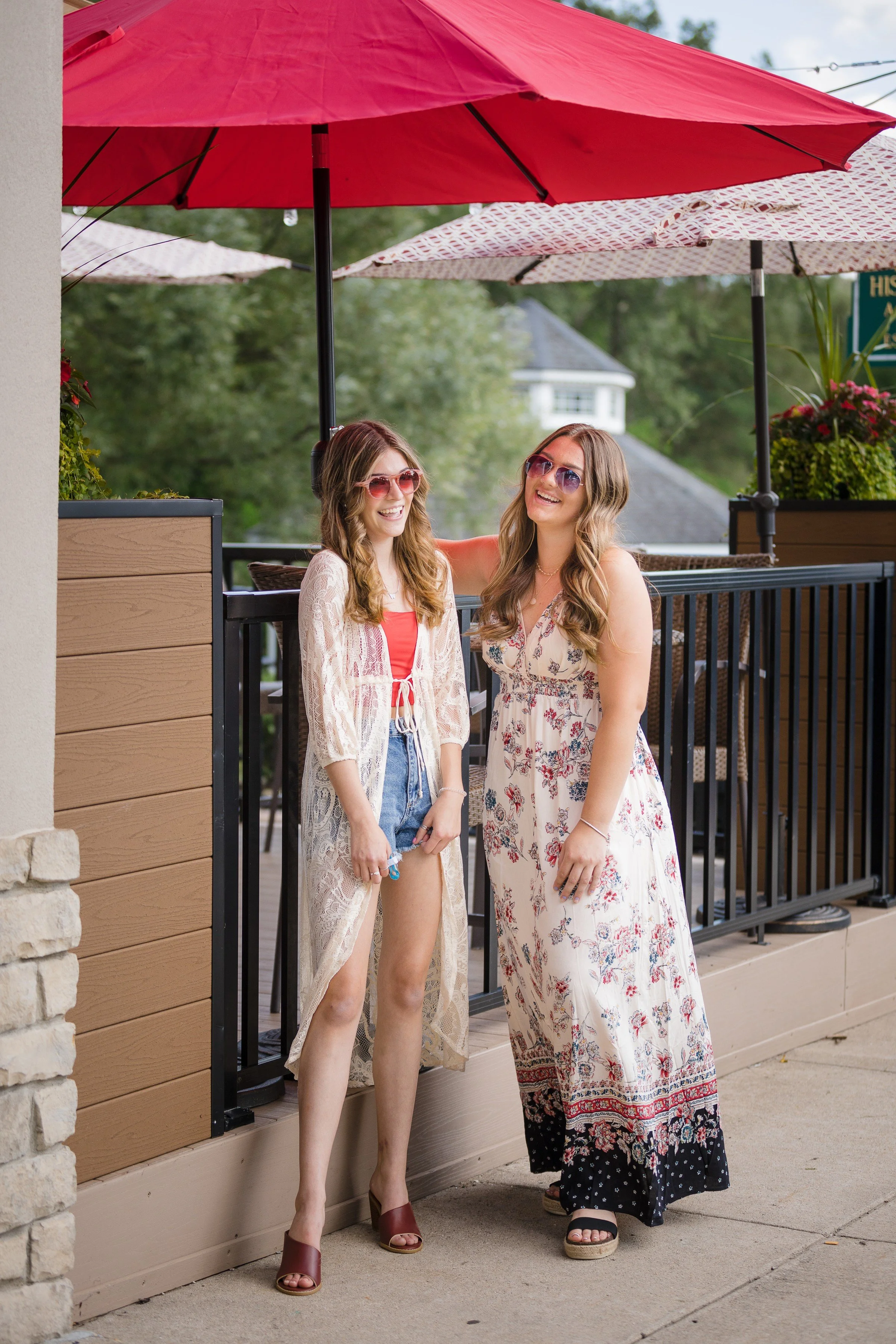Two women standing and smiling on a sidewalk under a large red patio umbrella. They are dressed in casual summer clothes, wearing sunglasses, and appear to be enjoying a sunny day outside.