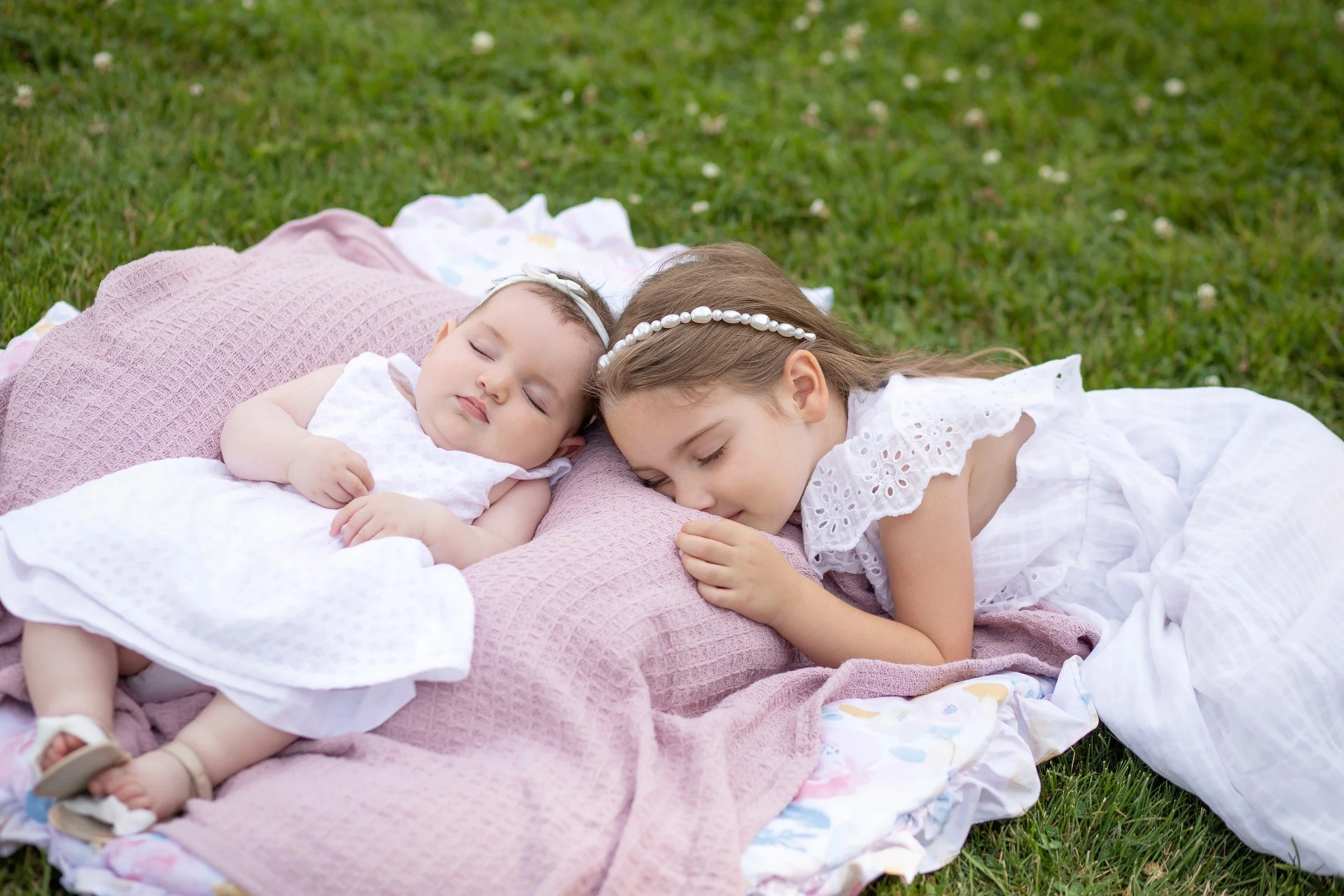 Two young girls sleeping on a pink and white blanket outdoors on the grass, one older and one younger.