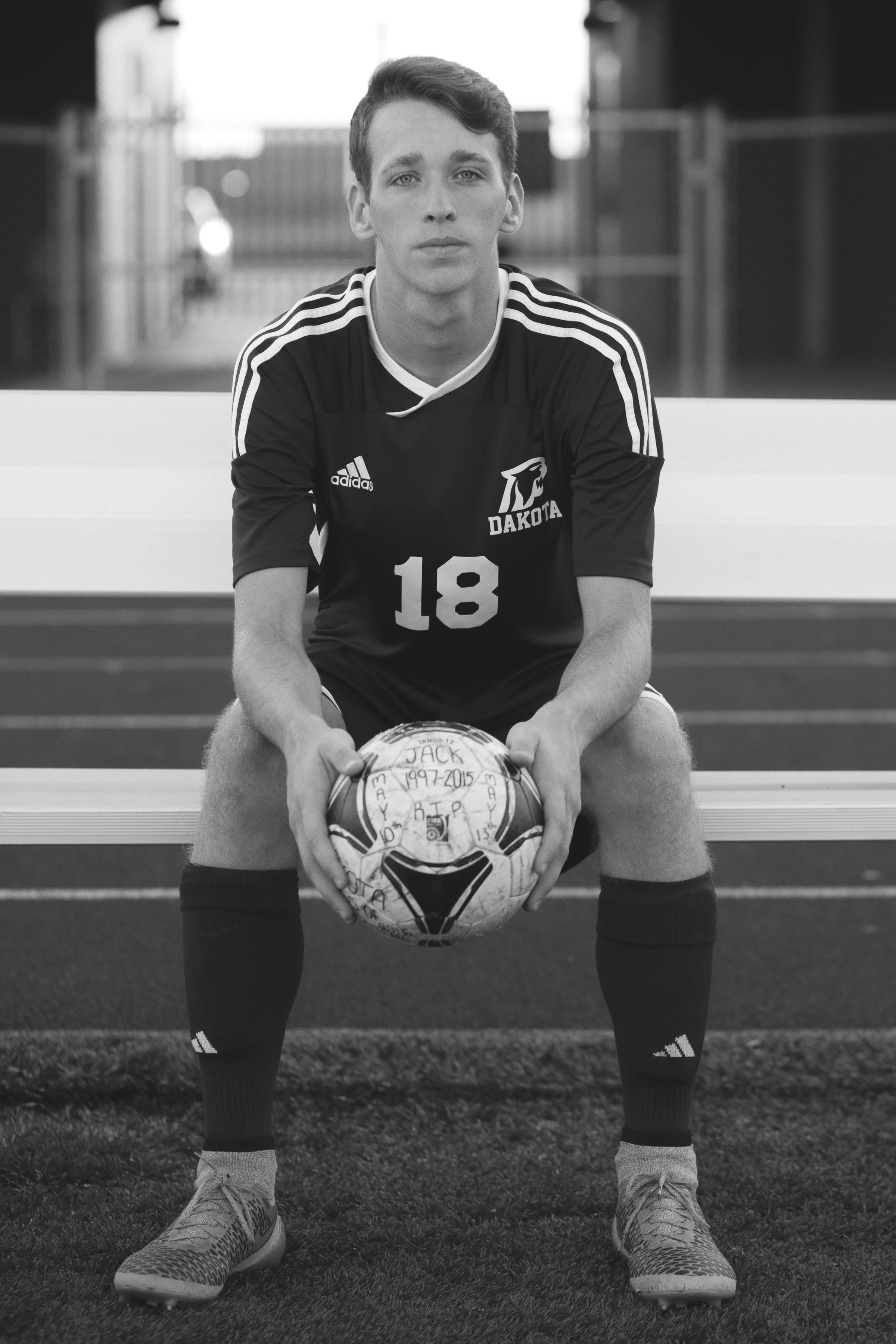 Young male soccer player sitting on a bench with a soccer ball, wearing a Dakota team jersey number 18, outdoors on a sports field.