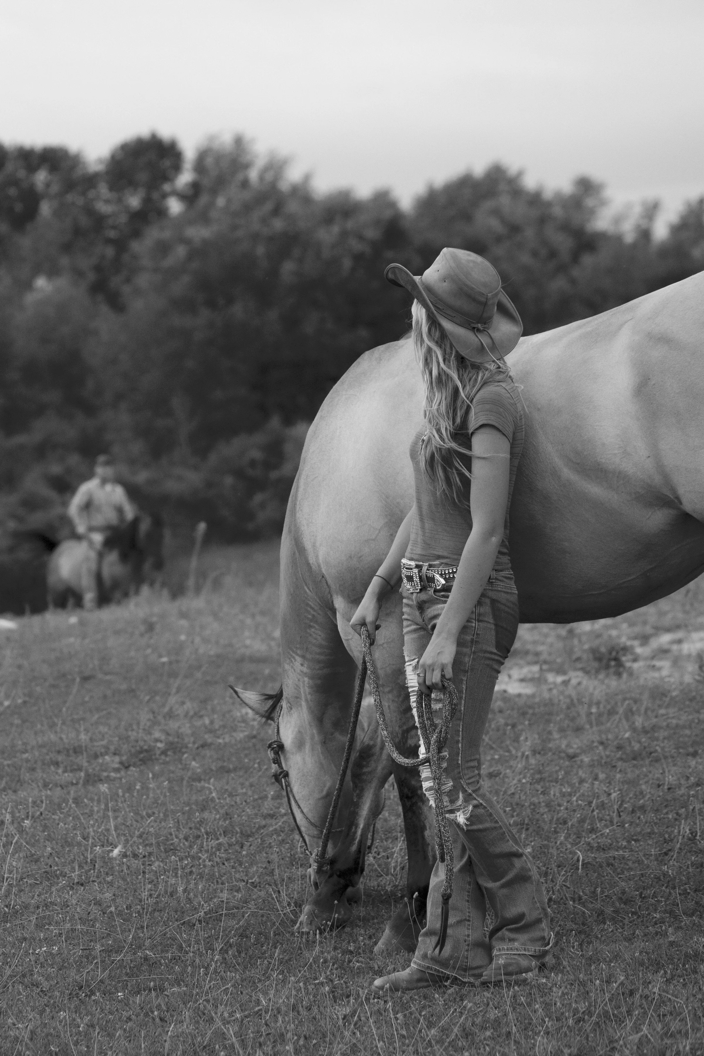 A young woman with long hair wearing a hat, t-shirt, and ripped jeans standing next to a horse while holding a halter. A person on horseback is visible in the background on a grassy field with trees.