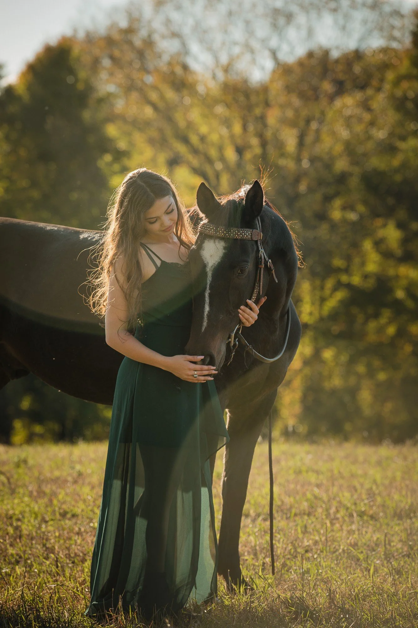 A young woman in a black dress standing in a grassy field, gently holding and leaning her head against a black and white horse with a decorative bridle, during sunset with a backdrop of foliage.