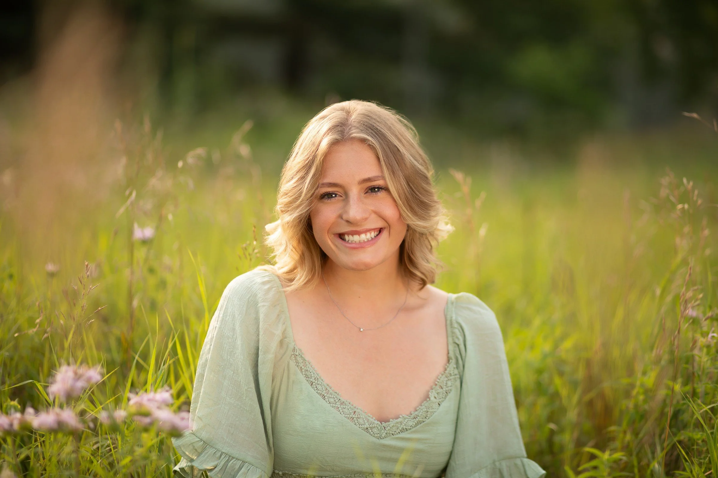 Smiling young woman with blonde hair sitting in a green field of tall grass and pink flowers, with a blurred natural background.