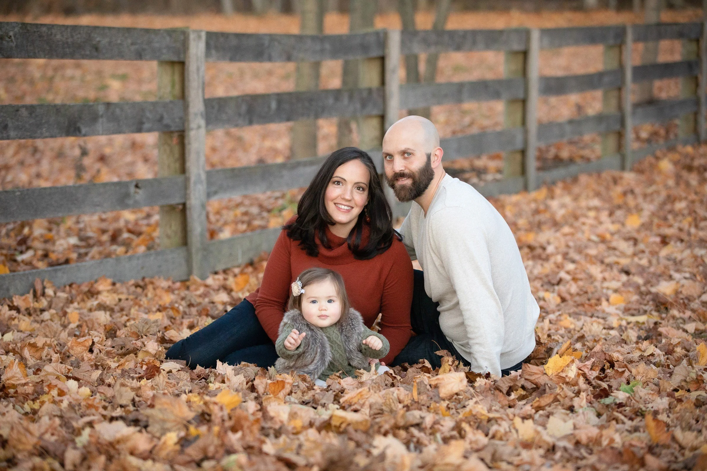 A family of three sitting on fallen autumn leaves in a park, with a wooden fence in the background. The woman has shoulder-length dark hair and is wearing a rust-colored sweater, the man has a beard and is bald, and the baby girl has light-colored ha