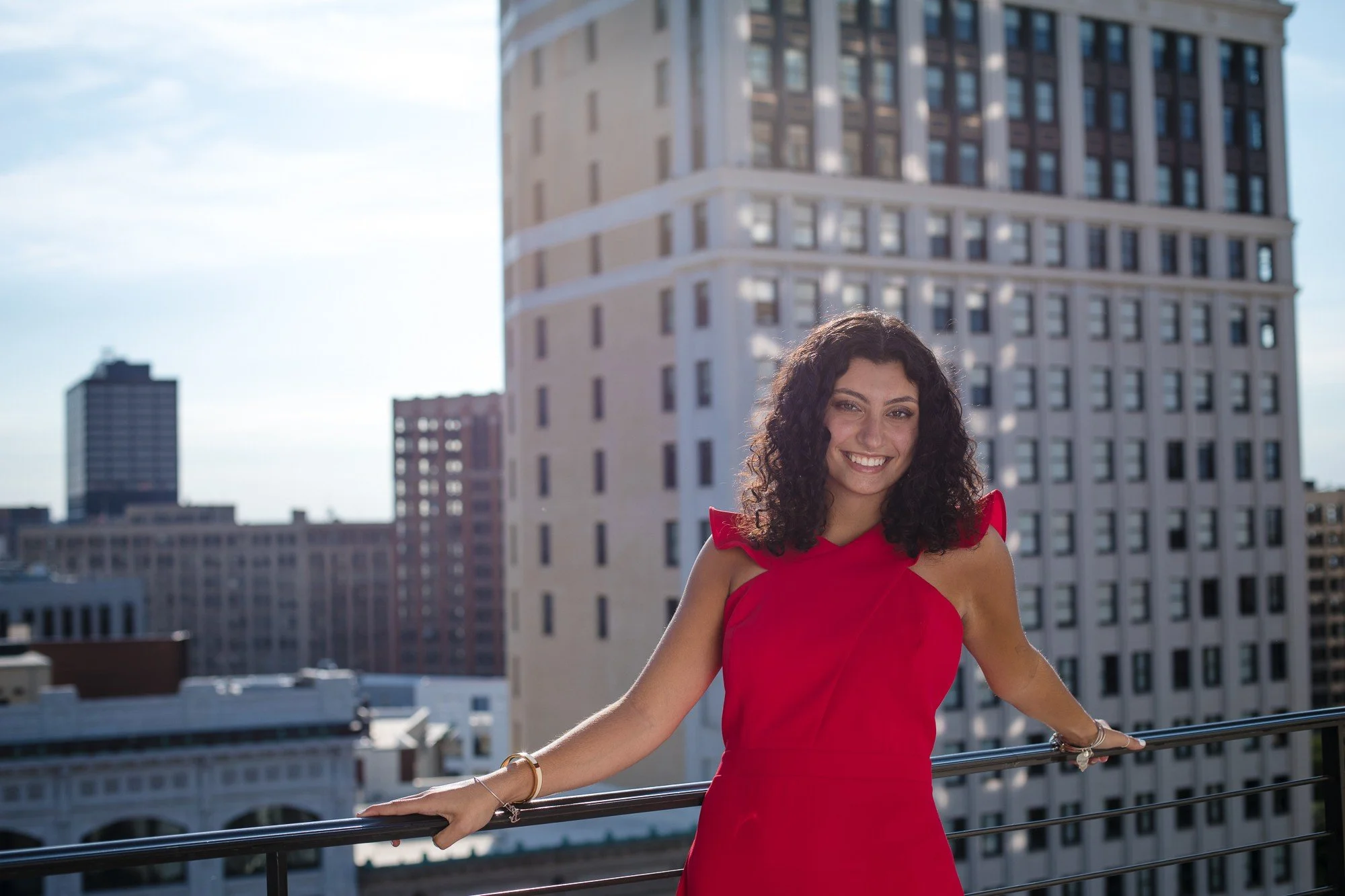 A young woman with curly black hair is smiling and wearing a red dress, standing on a balcony with a cityscape of tall buildings in the background.
