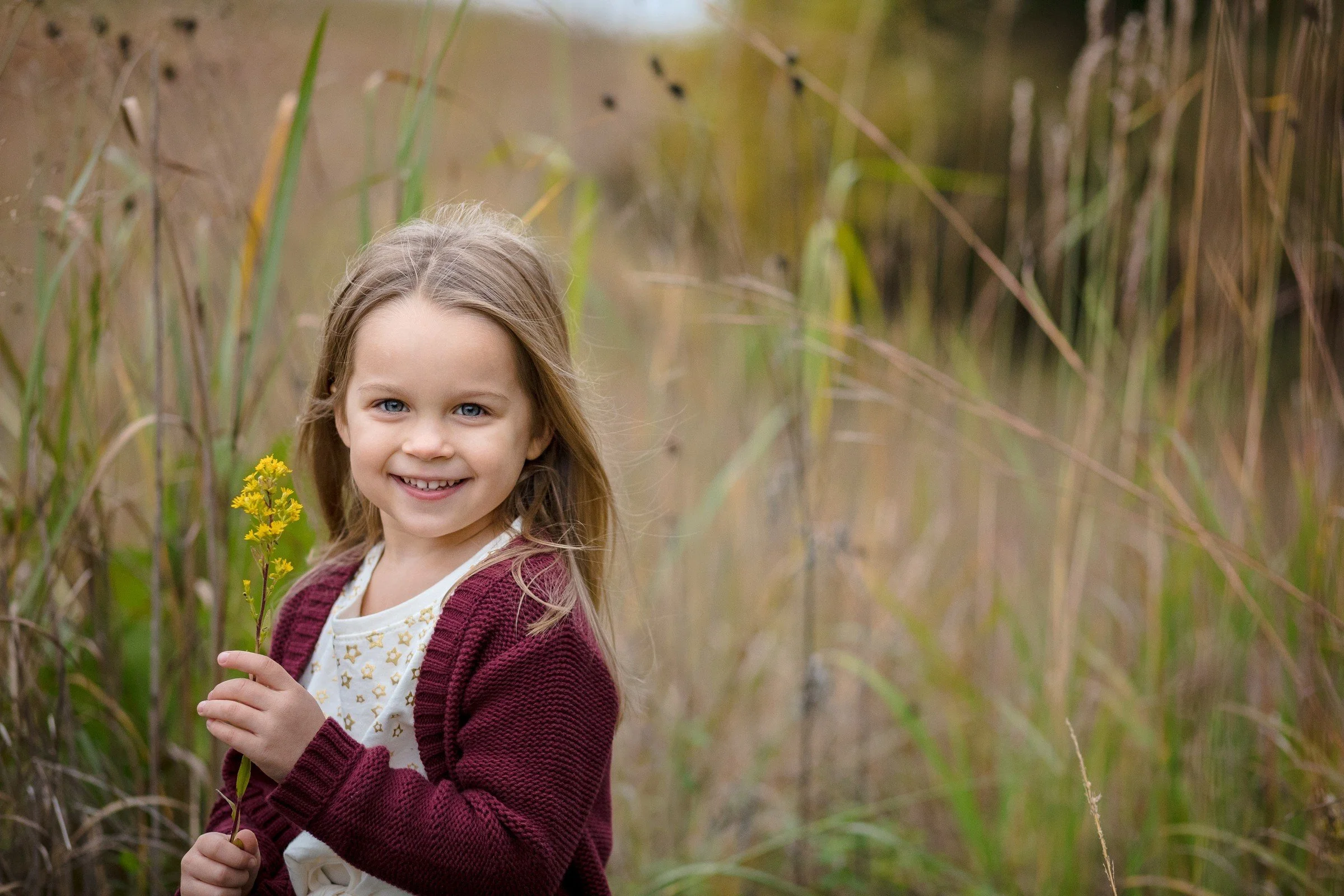 A young girl with long blonde hair, wearing a white shirt with gold stars, and a maroon cardigan, is smiling and holding a yellow flower in a grassy field.