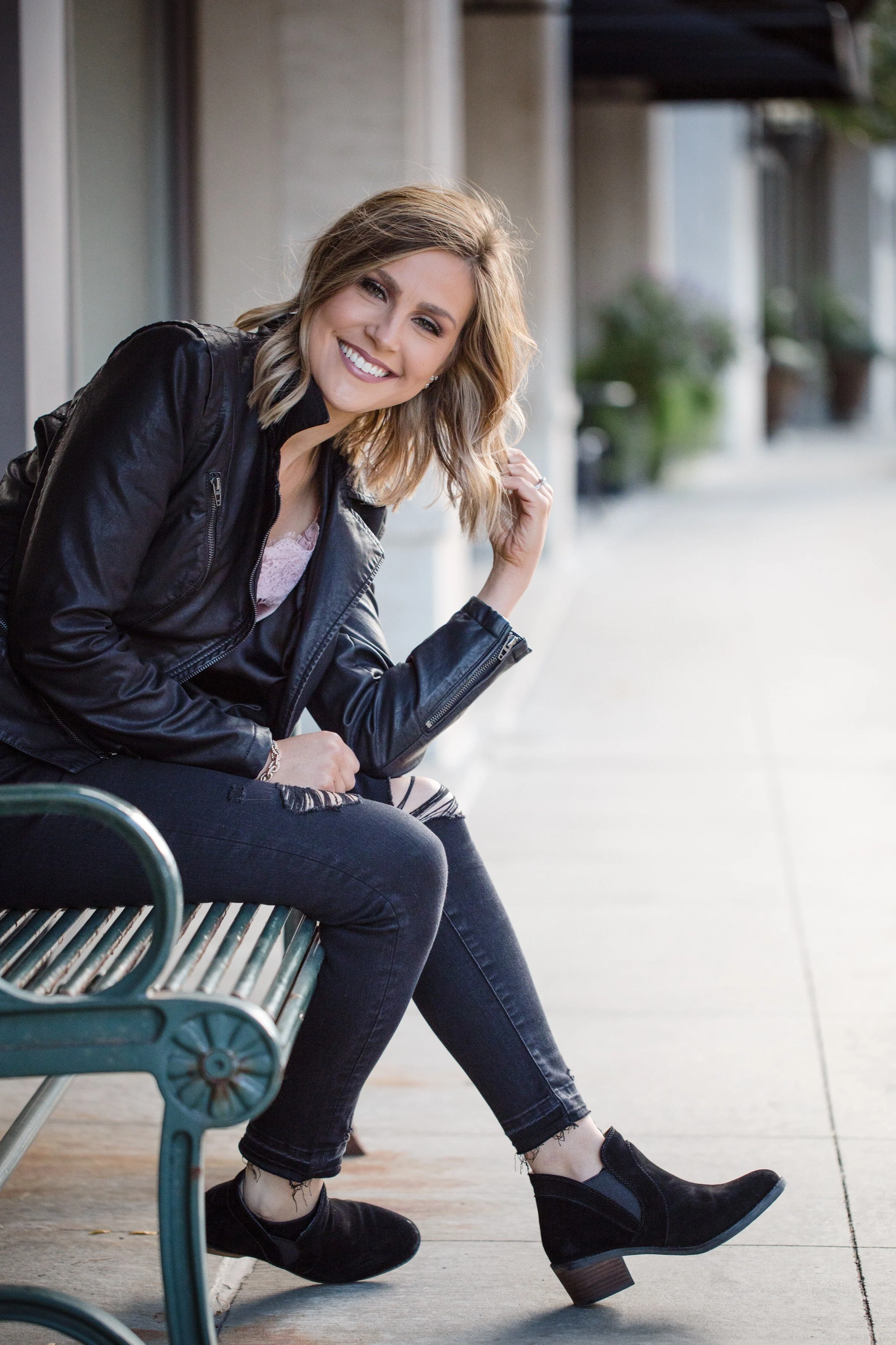 A woman sitting on a bench outside, smiling and looking at the camera. She has shoulder-length blonde hair, is wearing a black leather jacket, ripped black jeans, and black boots.