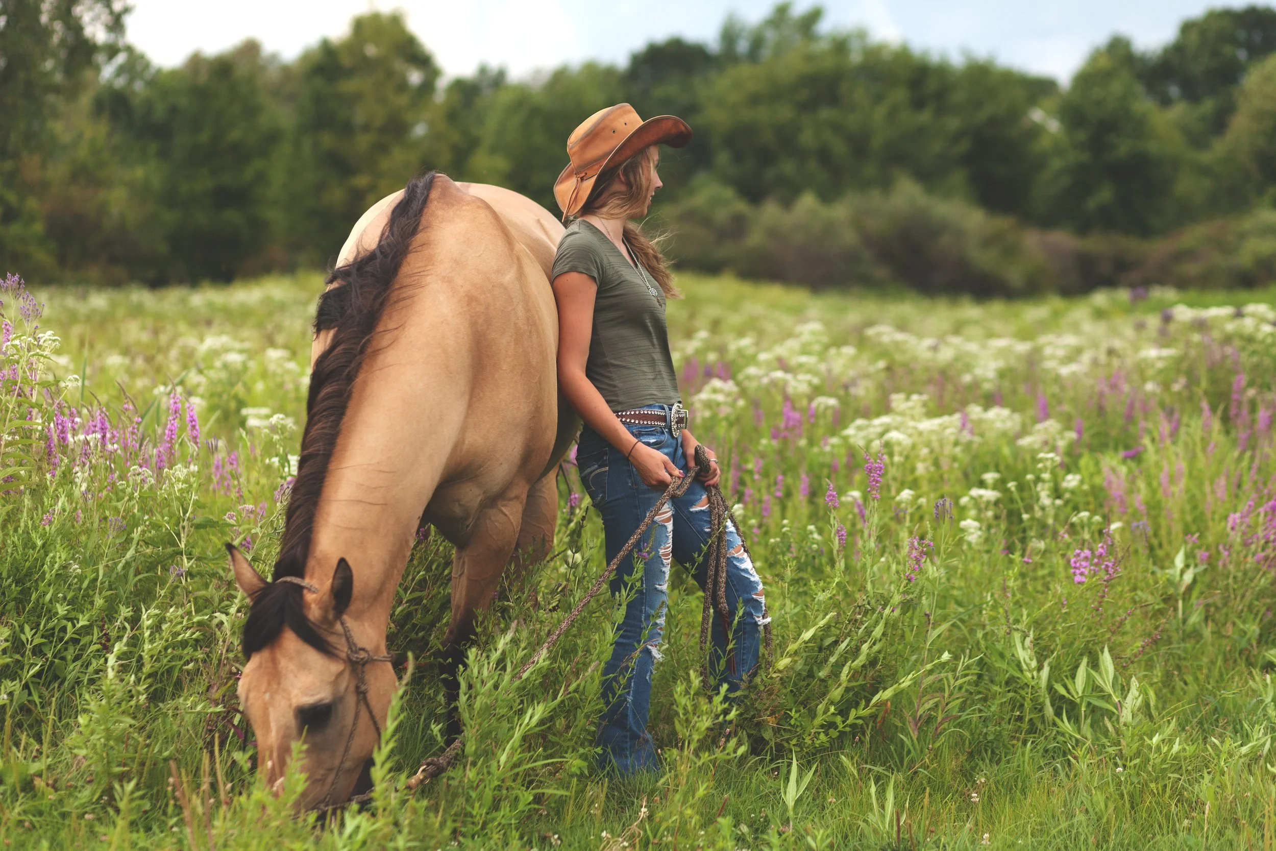 A woman in a cowboy hat standing next to a tan horse in a field of green grass and wildflowers.