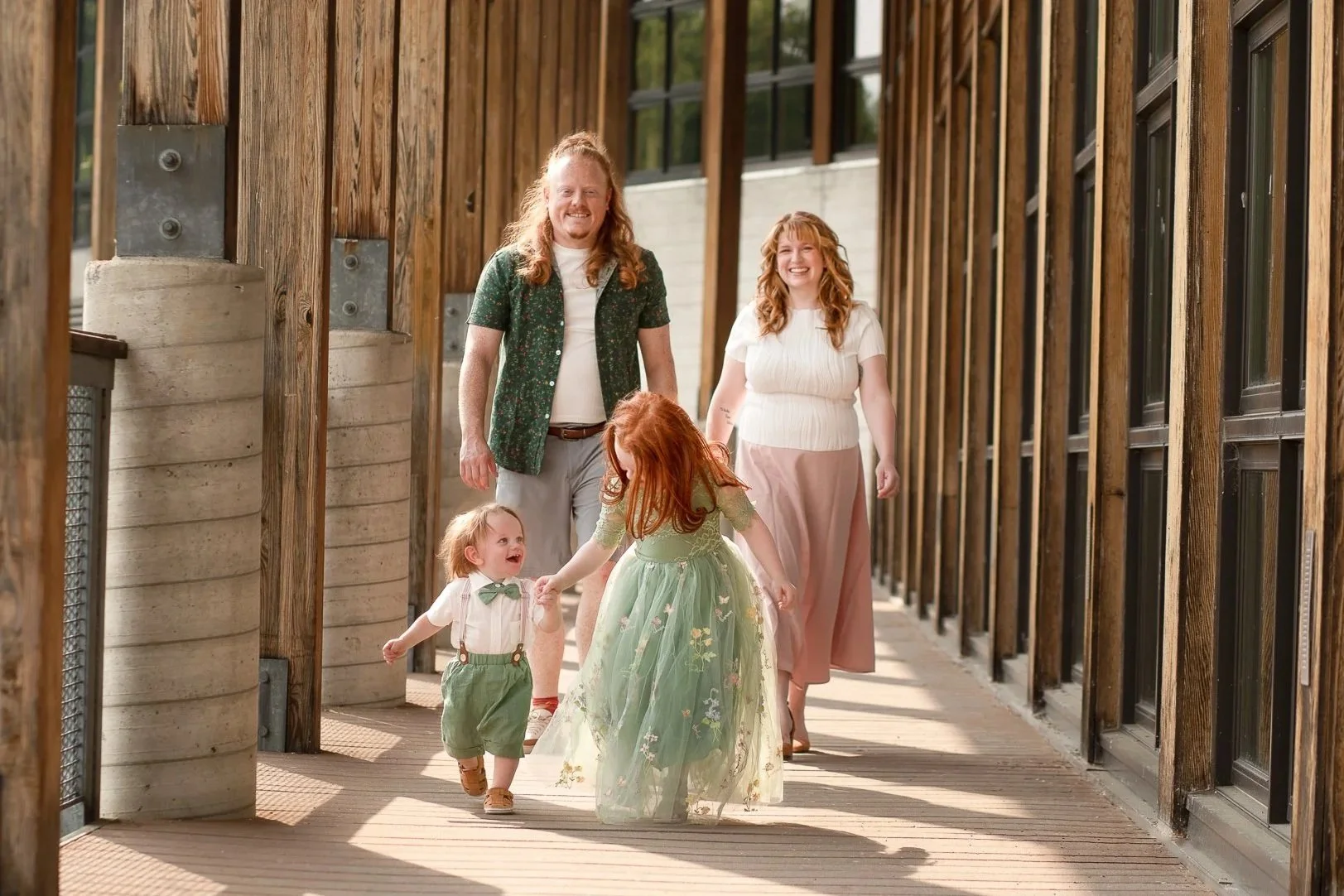A family of five walking on a wooden bridge outdoors, with two adults and three children, smiling and enjoying each other's company.