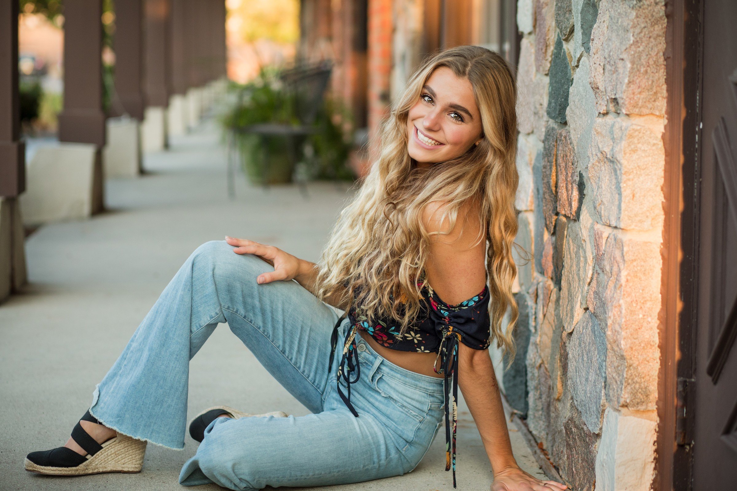 A young woman with long blonde hair smiling, sitting on the sidewalk against a stone wall, wearing a black floral crop top, light blue jeans, and black espadrille wedges.