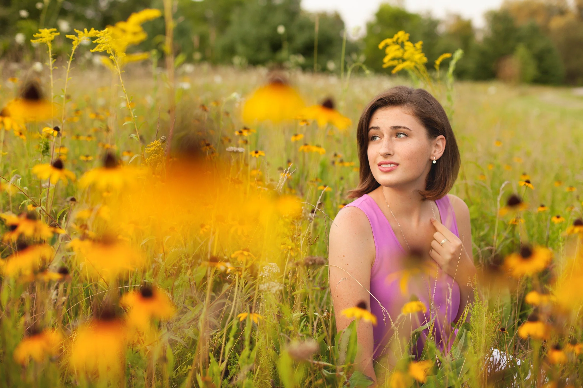 A young woman with short brown hair wearing a pink sleeveless top sitting in a field of yellow wildflowers, looking to her left.