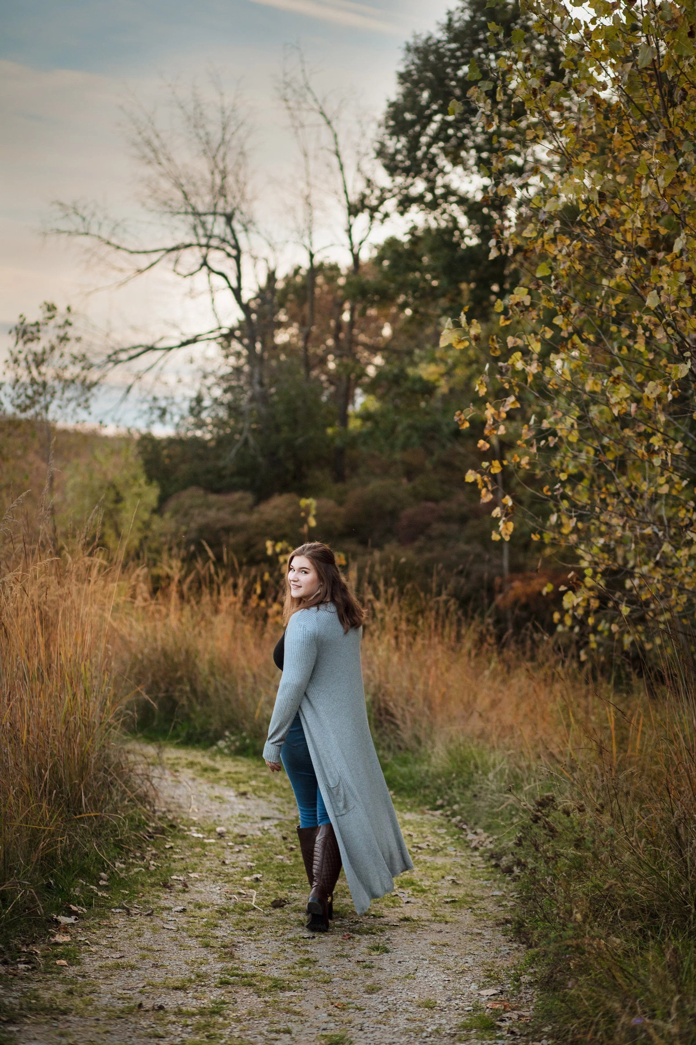 A young woman with brown hair, wearing a long light gray cardigan, black top, blue jeans, and brown knee-high boots, walking on a nature trail surrounded by tall grasses and trees during autumn, smiling and looking back.