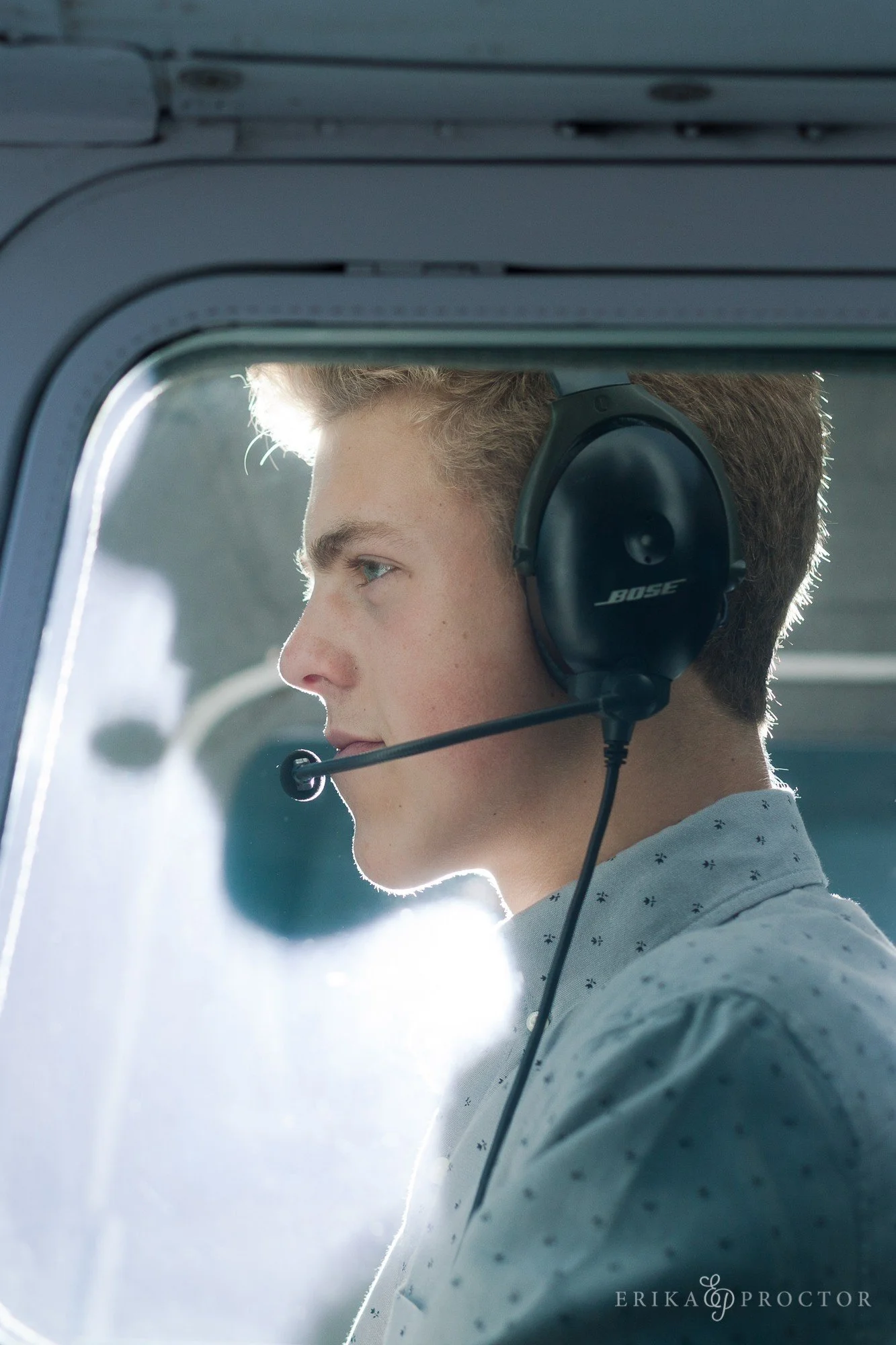 A young man with blonde hair wearing a headset with a microphone, sitting inside an aircraft cockpit or similar enclosed space, looking serious and focused.