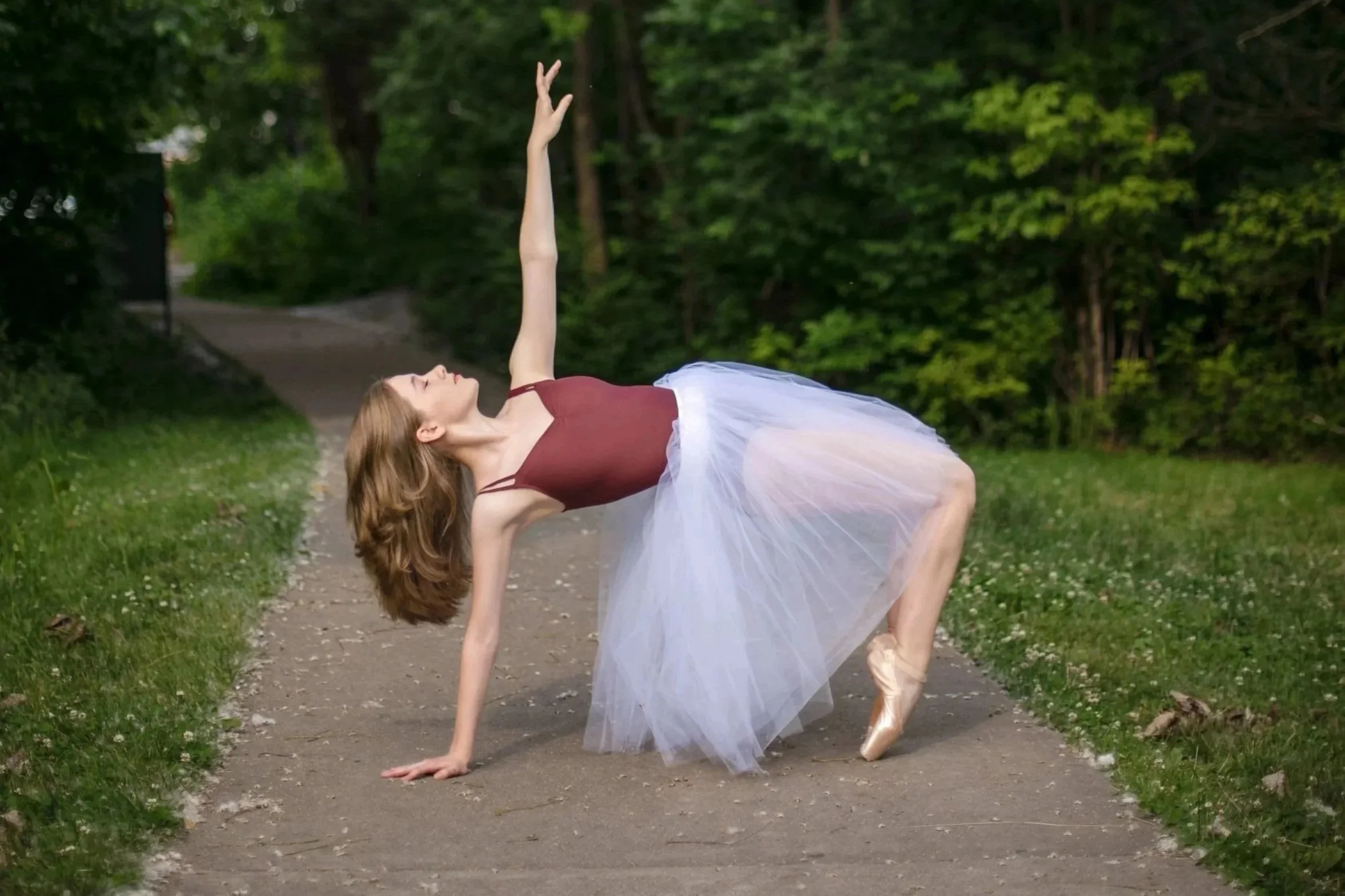 A ballerina performs a dance pose outdoors on a tree-lined path, wearing a maroon leotard and a white tutu.