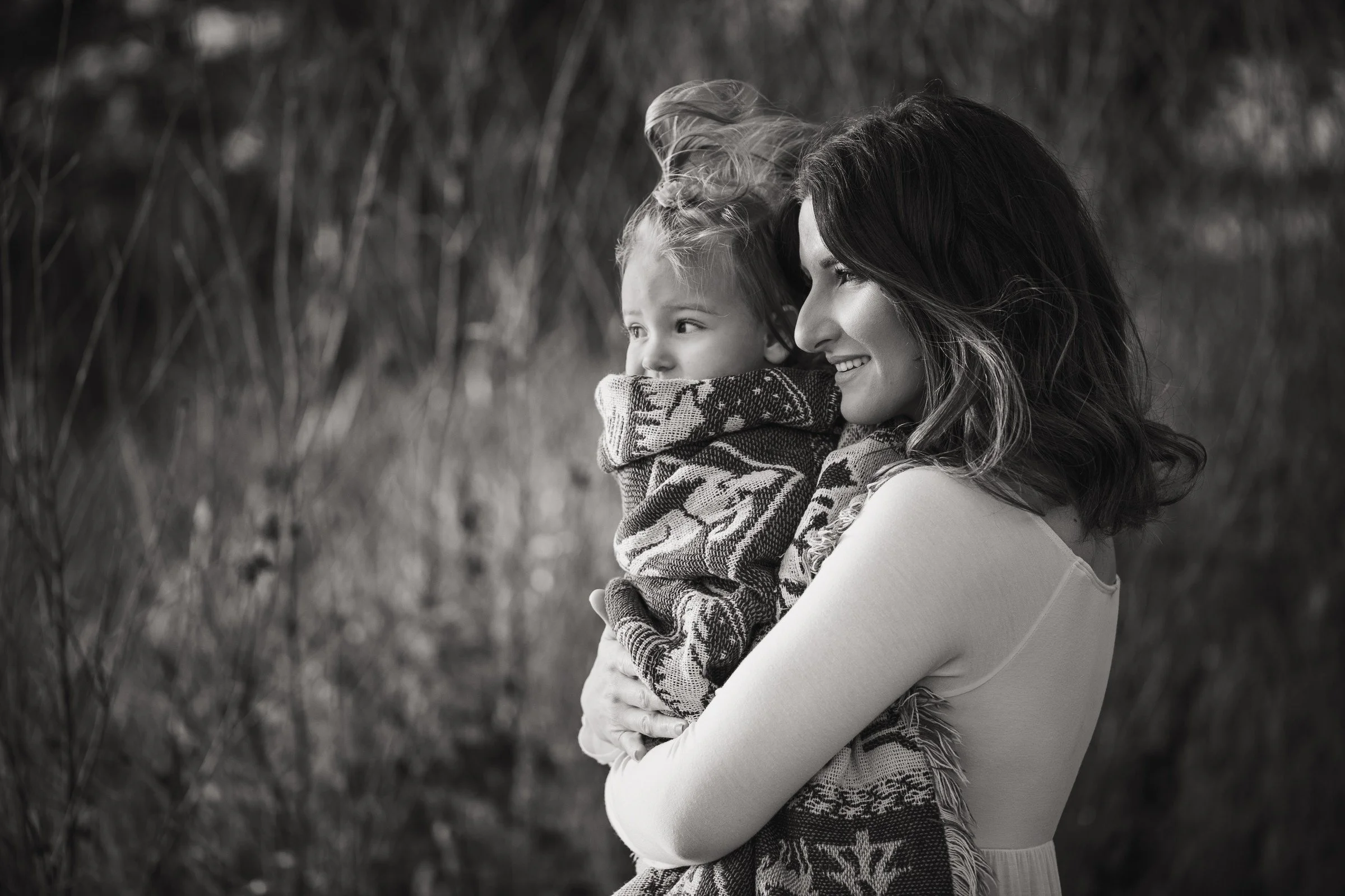 A woman holding a young child wrapped in a patterned blanket outdoors. They are smiling and looking into the distance.