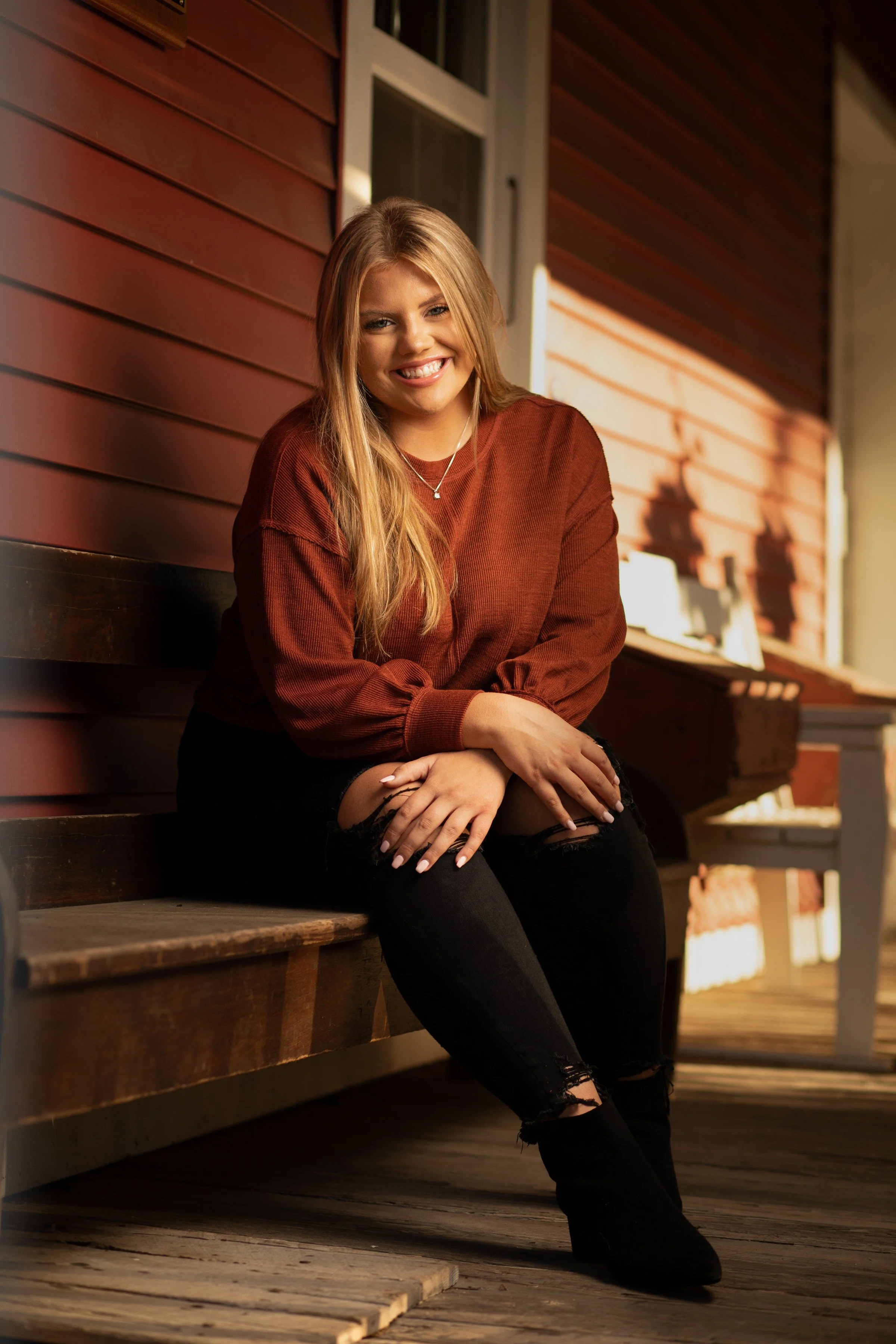 A young woman with long blonde hair smiling while sitting on a wooden bench outside a red house, wearing a maroon sweater and ripped black jeans.