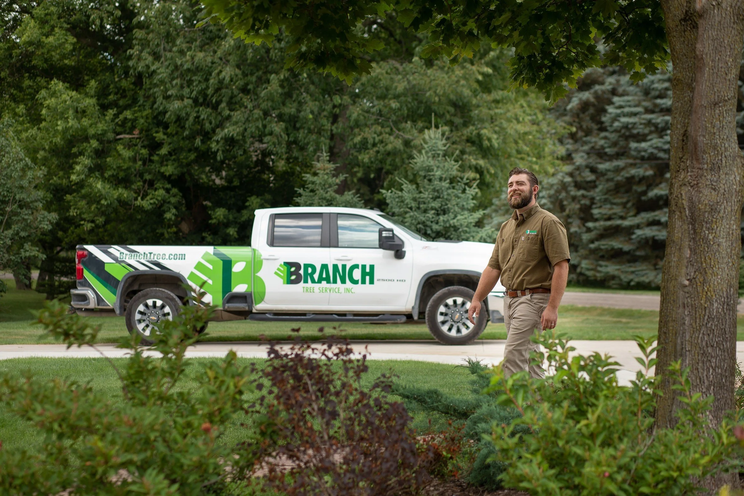 A man walking outdoors near a tree, with a company truck in the background labeled 'Branch Tree Service, Inc.' and a website URL 'BranchTree.com.' The man has a beard and is wearing a brown shirt and khaki pants, smiling as he walks on a sidewalk.